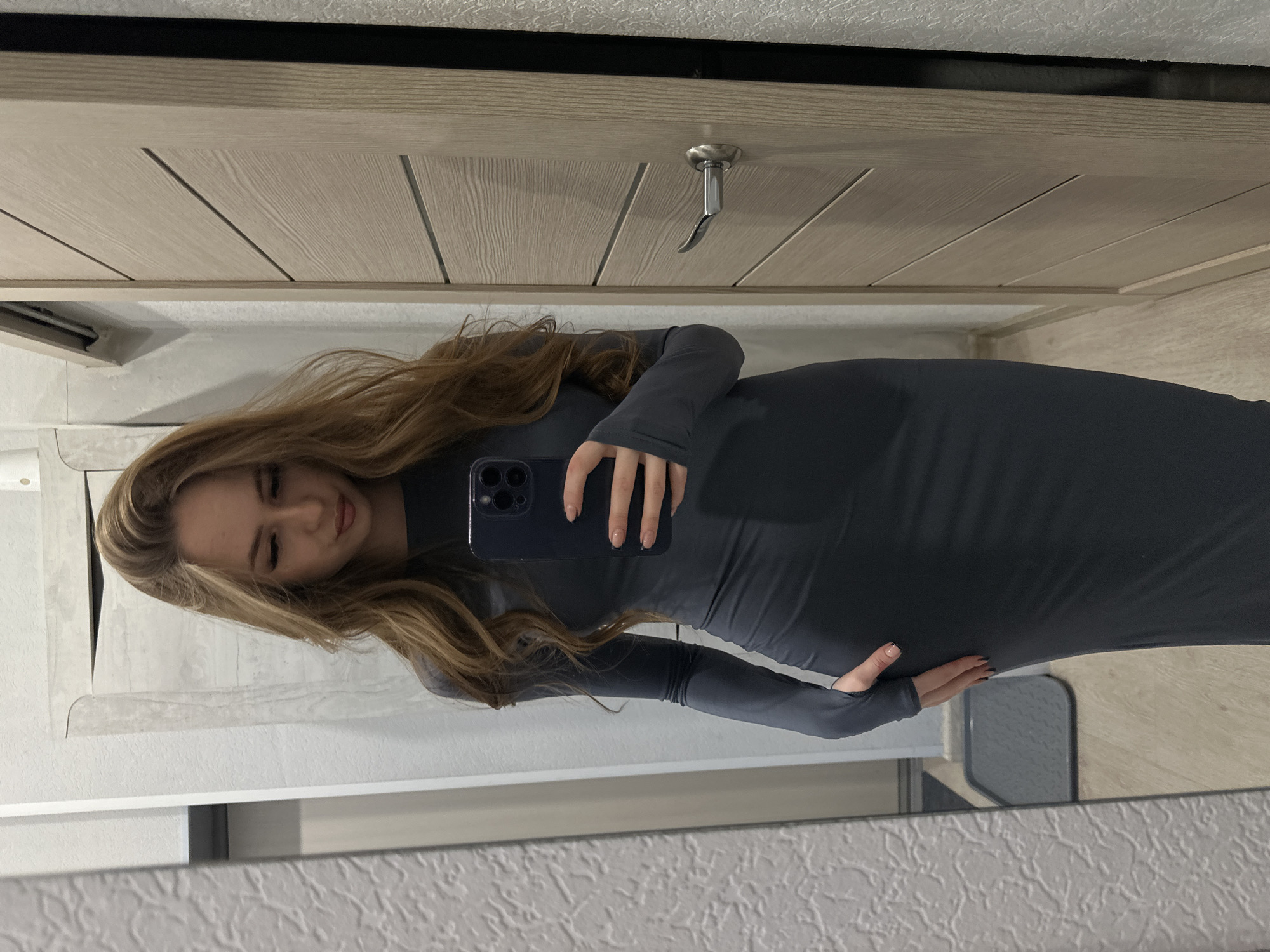A woman is posing in a mirror in an indoor setting, wearing a fitted gray dress with long sleeves. She is holding a dark-colored smartphone, presumably taking a selfie. Her hair is long, wavy, and light brown. The background includes a light-colored, panelled door with a silver handle. The lower portion of the image shows a mirror frame and a glimpse of the room's decor. The location is not specified, as the city and area are unknown.