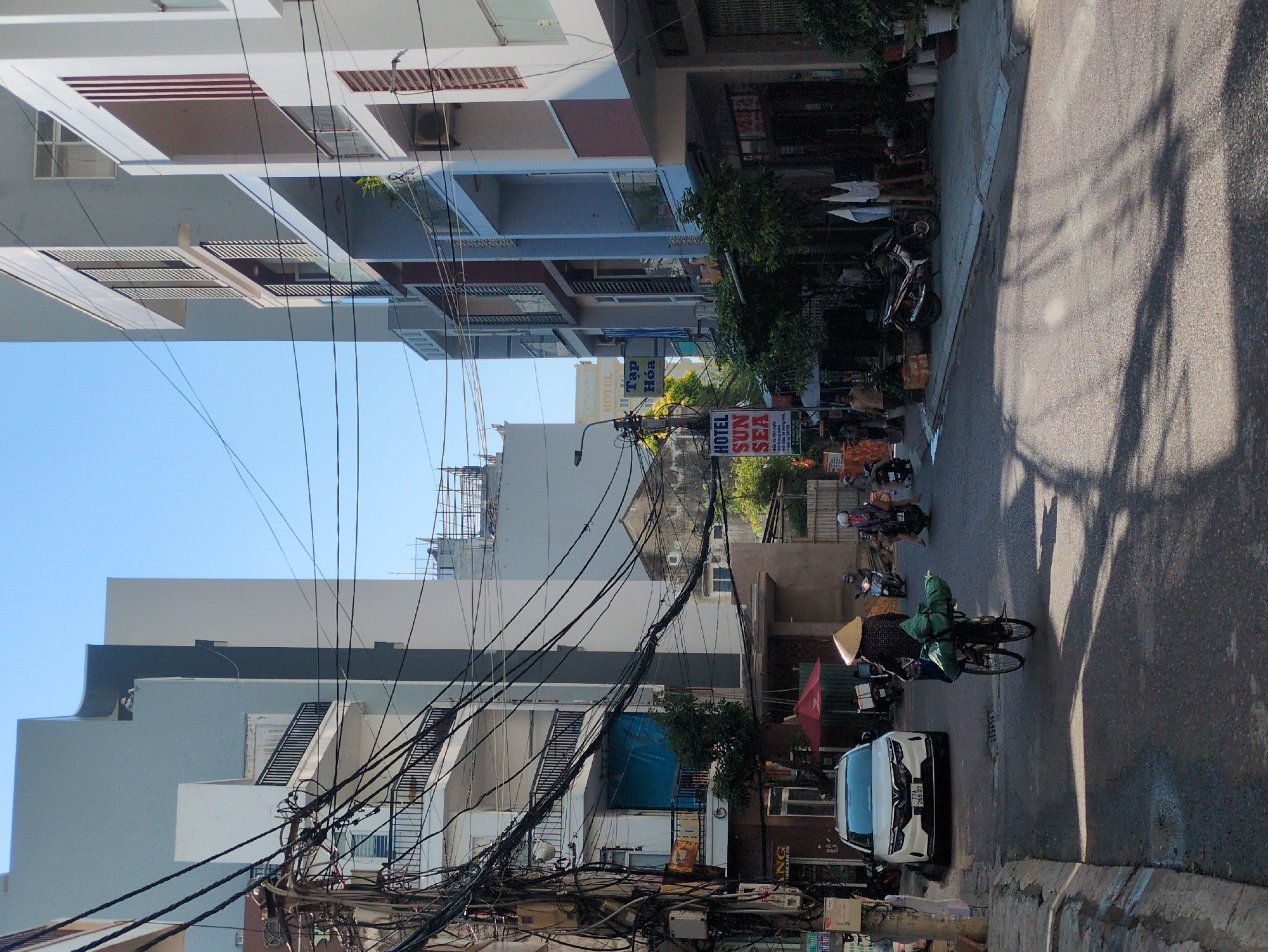 This image captures an urban street scene in South Quy Nhon Ward, Vietnam, under a clear blue sky. The paved asphalt road shows prominent, elongated shadows indicating strong overhead sunlight.
In the foreground, a person wearing a conical hat and dark clothing is riding a bicycle away from the viewer, positioned centrally in the lane. A large green bundle is secured to the rear rack of the bicycle. To the left, a white SUV with license plate 77A-269.07 is parked parallel to the sidewalk. On the far left, a utility pole is heavily laden with dense, black electrical and communication cables.
Further down the street, multiple motorcycles are visible; two individuals are riding one, while others are parked along the right-hand sidewalk. Buildings line both sides of the street. On the left, multi-story structures feature dark-railed balconies and partially visible ground-floor shopfronts, some obscured by the utility wiring. On the right, newer, multi-story buildings display a mix of modern architecture with balconies, some featuring glass railings and others with vertical louvred elements. A prominent red and white sign for "HOTEL SUN SEA" is visible, along with a "Tạp Hóa" (convenience store) sign. Potted plants and other street furniture are present on the sidewalks.
In the background, taller, lighter-colored buildings rise against the horizon, with some rooftop structures visible on the most distant buildings.