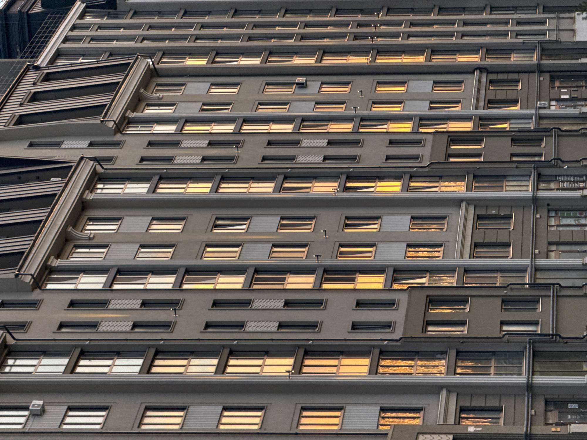 The image shows a close-up of a building facade. The building is gray with numerous windows arranged in horizontal rows. Sunlight is reflecting off the windows, creating a golden glow. There are metal structures and pipes running vertically along the building's exterior. The architectural style appears modern, but there is no indication of a specific location in Zelenogradsk, Russia, or any other city. No people are visible in the image.