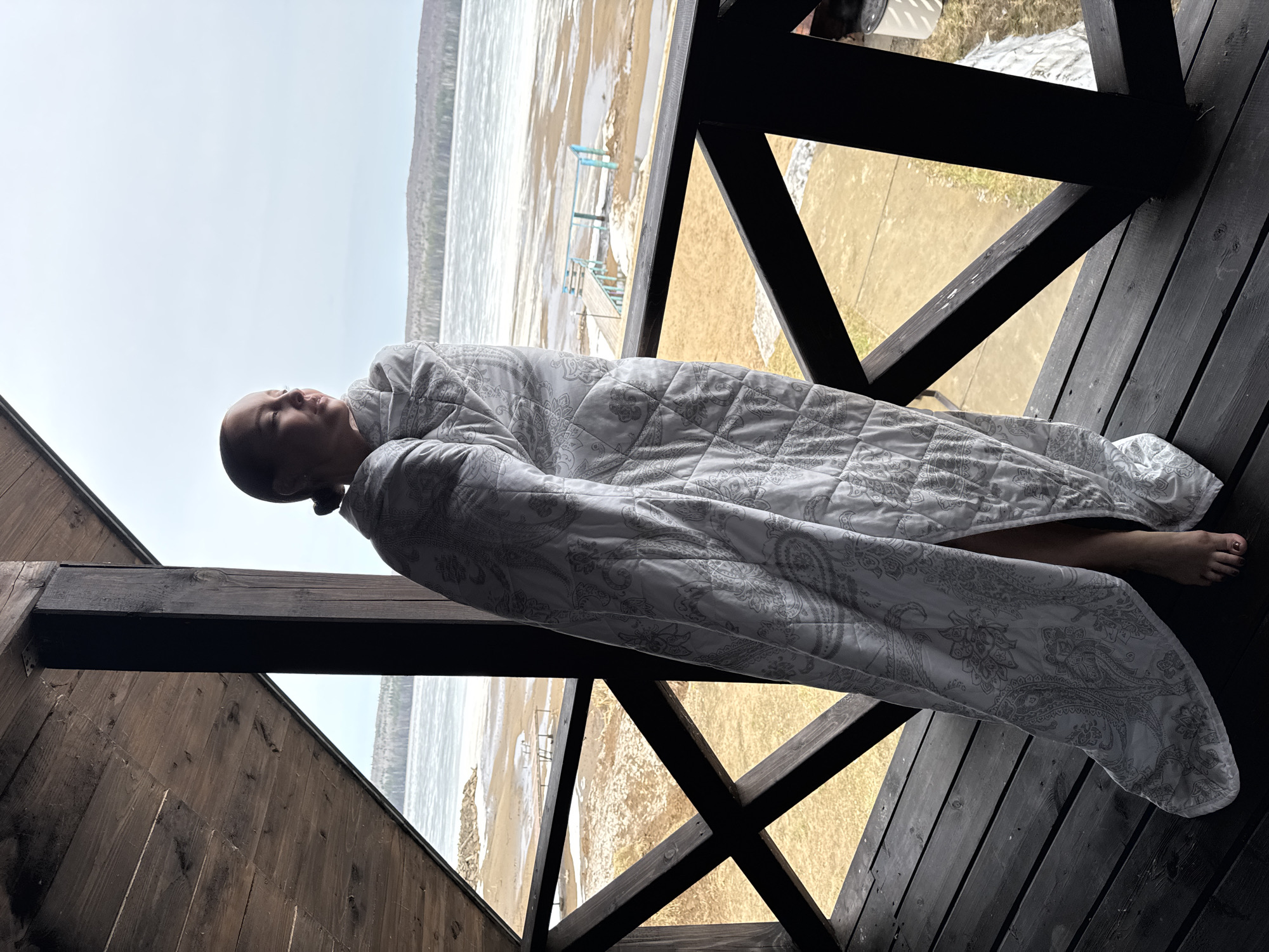 A person is lying horizontally, fully wrapped in a white patterned quilt, on a wooden deck. The deck has a railing of dark-colored wooden planks arranged in an X-shape pattern. The person is facing left, and only their face is visible above the quilt. A single bare foot extends from the bottom of the quilt.
Beyond the deck, a partially frozen body of water stretches towards the horizon, with what appears to be land or low hills in the background. A beach is visible between the deck and the frozen water. There are small structures on the beach. The sky is overcast and grey.
Based on visual elements, particularly the presence of a frozen body of water, the location is most likely outside, during a cold season. The environment includes natural elements like the water, beach, and sky, as well as man-made structures like the deck, railing, and buildings in the background.