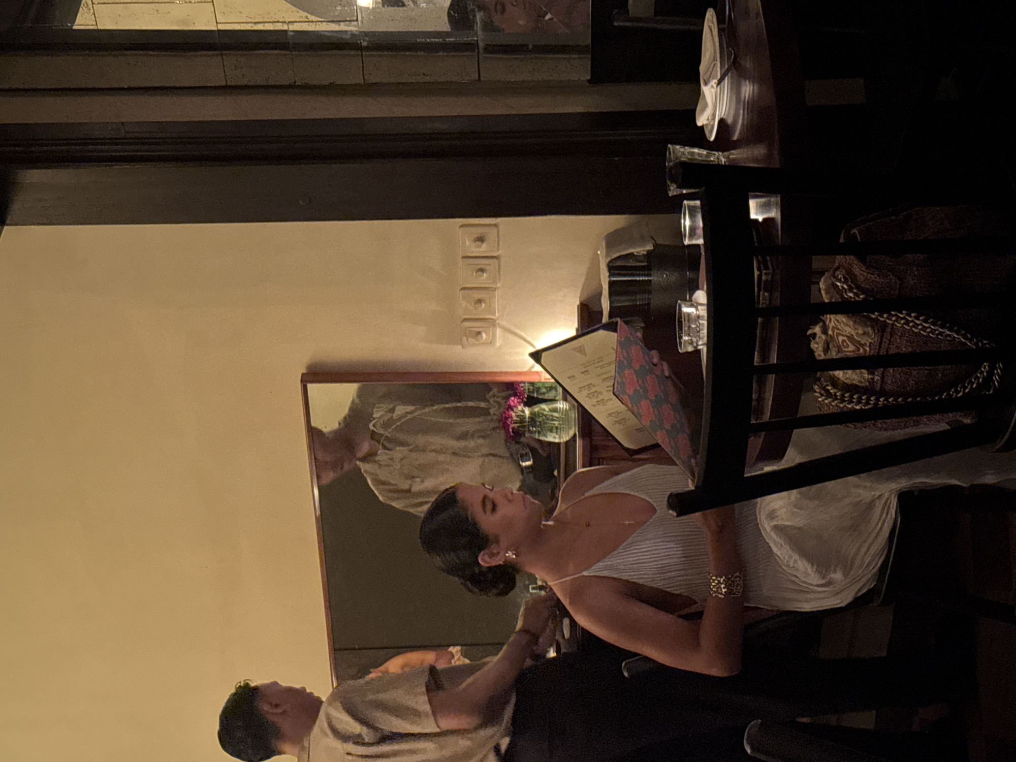 An indoor scene depicts a couple having dinner in a dimly lit restaurant or dining establishment, likely in the evening. The image is oriented vertically (originally rotated 90 degrees clockwise).
At the center, a woman is seated at a dark wooden table, viewed from her side. She wears a light-colored, possibly striped, spaghetti-strap top or dress, and her dark hair is styled in a bun. She wears small earrings and a prominent gold-colored cuff bracelet on her left wrist. Her gaze is directed downwards at a patterned menu she holds in her hands. To her right, another dark menu with faint white text rests on the table.
Opposite her, a man is partially visible, seated at the same table. He wears a light-colored (beige or grey) short-sleeved shirt. His reflection is clearly visible in a large rectangular mirror behind the woman. The mirror also reflects a small vase containing purple or pink flowers, positioned on a surface behind the table.
The setting features light-colored walls (beige or off-white) and dark wooden beams or trim running across the top portion of the frame. On the wall, a stack of four white square light switches or outlets is visible. Several clear drinking glasses, some appearing to contain liquids, are placed on the table. Behind the woman, on a dark chair or bench, a patterned fabric with a chain-like design is visible. Stacked white plates or bowls can be seen on a shelf or surface in the background.
The overall atmosphere is intimate and cozy, characteristic of a dinner setting. No legible text is visible in the image. Given the location context of Mengwi, Indonesia, the restaurant likely incorporates local design elements.