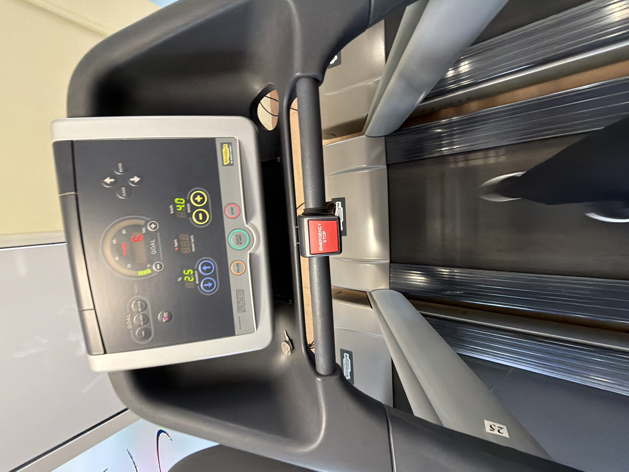 The image shows the console and handrails of a Technogym treadmill, with a partial view of a person's leg and foot on the treadmill belt. The treadmill console displays speed, gradient, and goal metrics. An "Emergency Stop" button is visible on the handrail. The environment appears to be indoors, likely a gym or exercise facility.