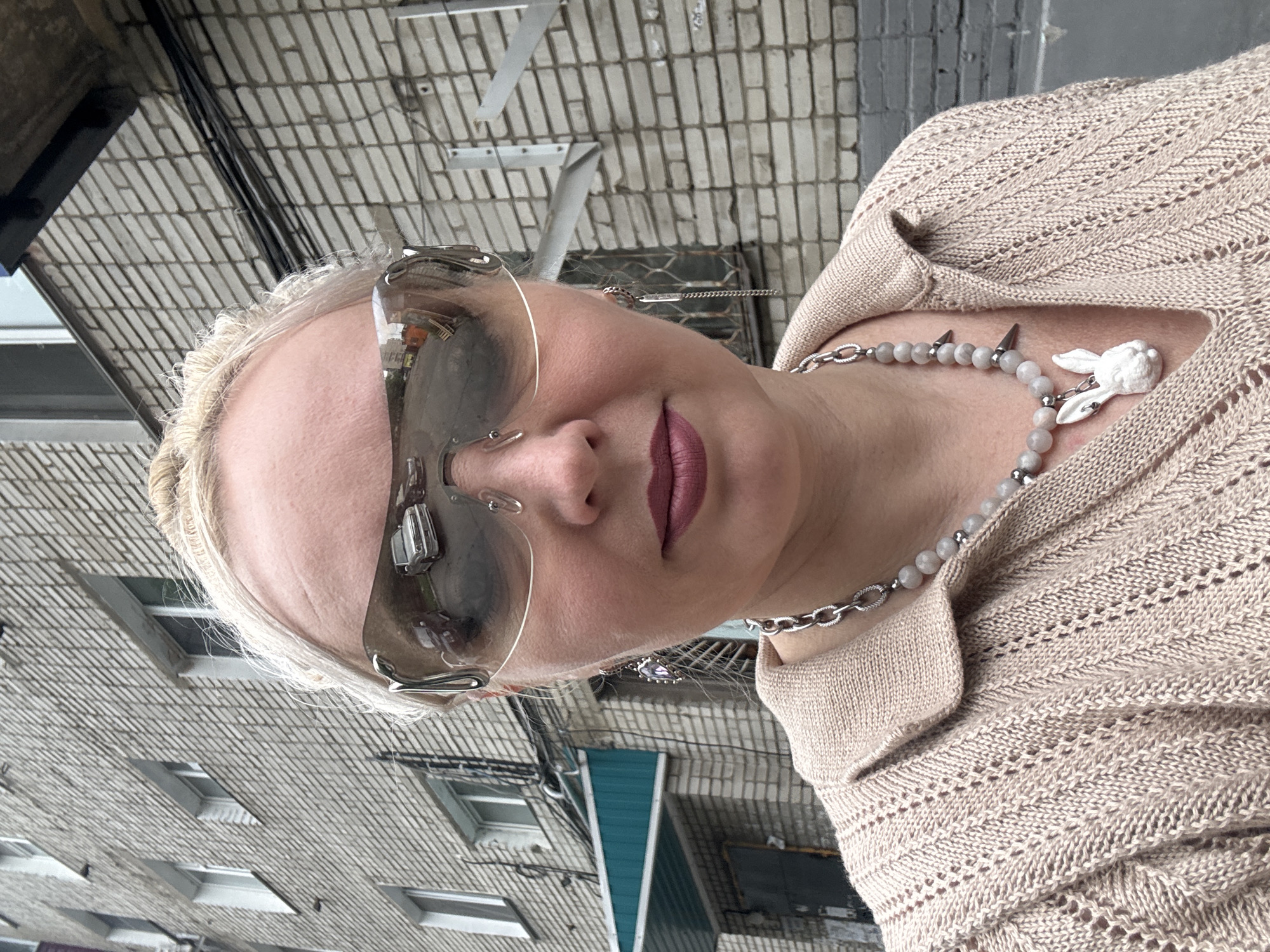 A blonde woman wearing sunglasses, a beige knit sweater, a silver chain necklace, and a white bunny pendant stands in front of a brick building in Komsomolsk-on-Amur, Russia. The sunglasses reflect a street scene with cars. The building appears to be a residential or industrial structure, with visible windows, electrical conduits, and metal brackets on the exterior. The lighting suggests an overcast or shaded daytime environment.