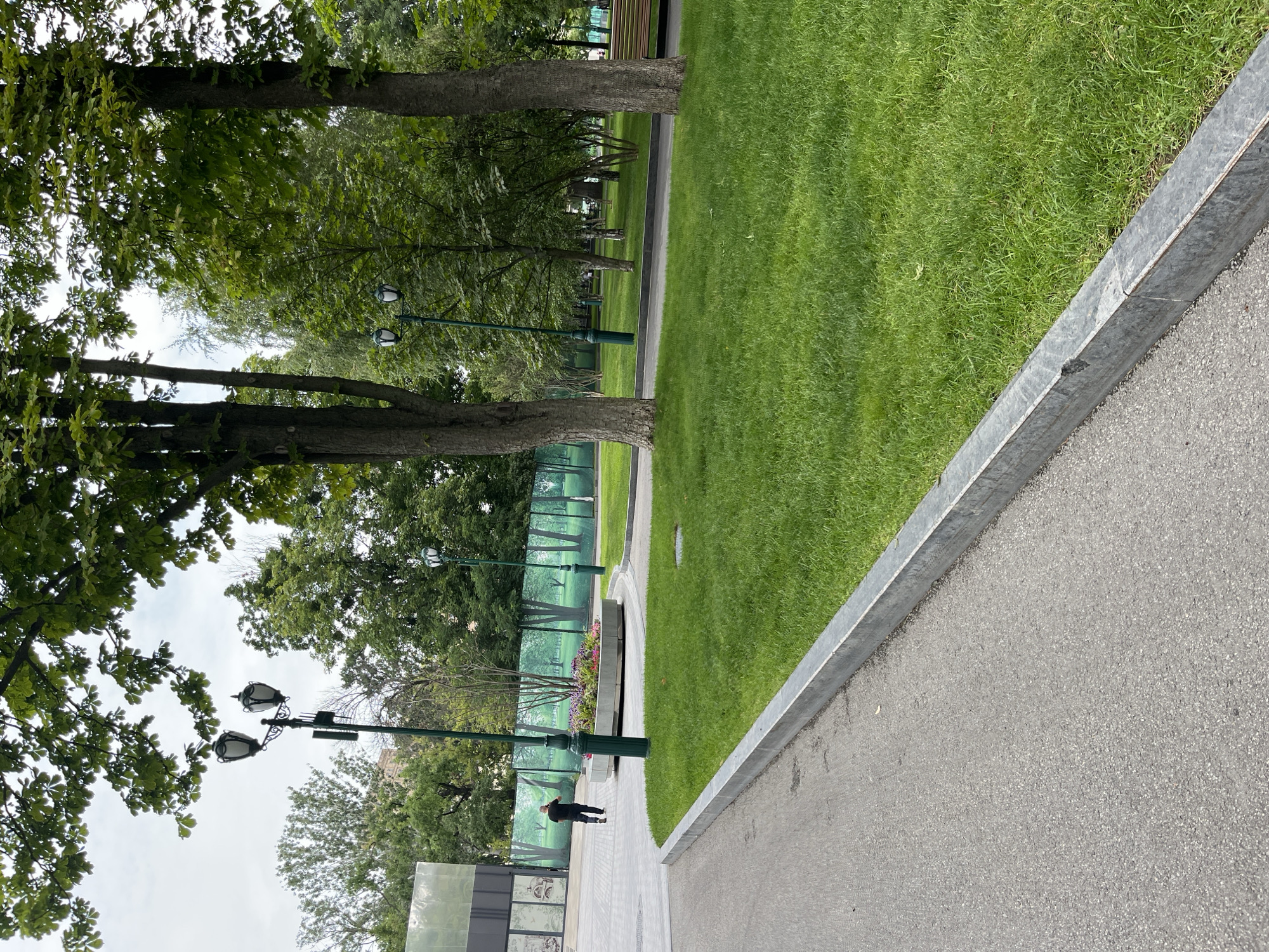 The image depicts a park scene in Kharkiv, Ukraine. In the foreground, a grey asphalt path curves to the left, bordered by a metal curb. To the right of the path is a well-maintained green lawn. Further back, a circular flower bed filled with colorful flowers is visible. Several tall trees with green foliage are scattered throughout the park, casting shadows on the lawn. Two ornate street lamps with double lights stand in the background. A man in a black shirt and dark pants is walking away from the camera on the paved area. A large, modern building with a glass facade is visible on the far left. A green fence with a printed mural of trees runs along the middle ground, creating a backdrop for the park. The sky is overcast.