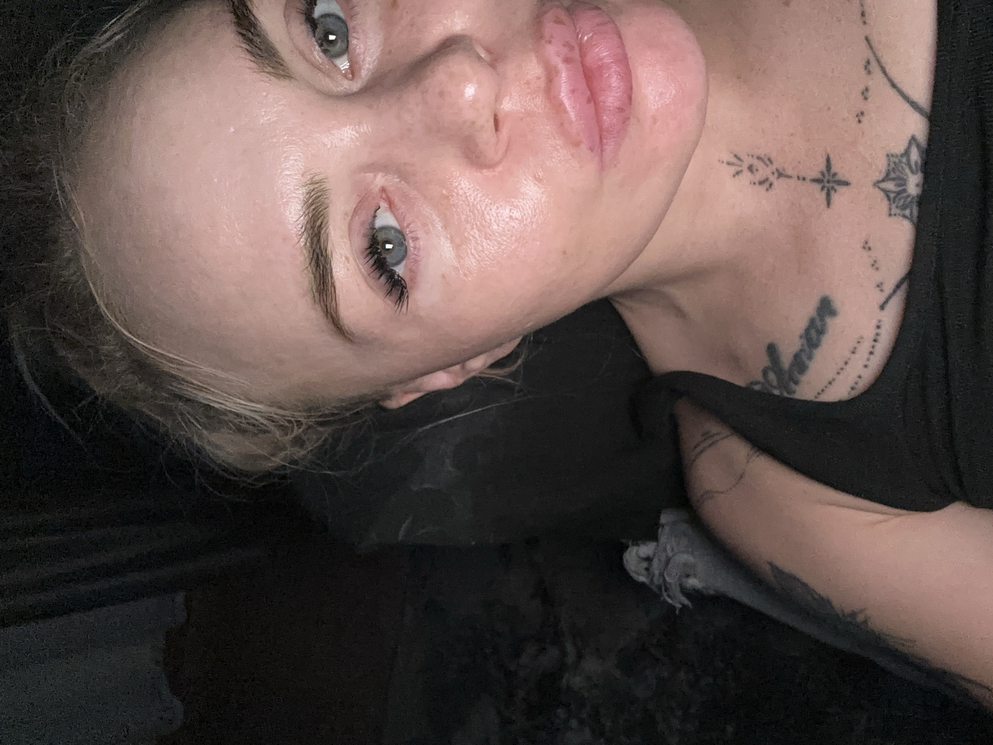 A close-up, waist-up portrait of a blonde woman with pale skin, freckles, and blue eyes is shown. She is wearing a black tank top with tattoos visible on her chest and left arm. The woman's mouth is pursed in a kiss-like gesture. The background is dark and out of focus, with a hint of black fabric to the left. The lighting appears to be artificial, creating a slight sheen on her skin. The image is taken from a slightly high angle, looking down at the woman.