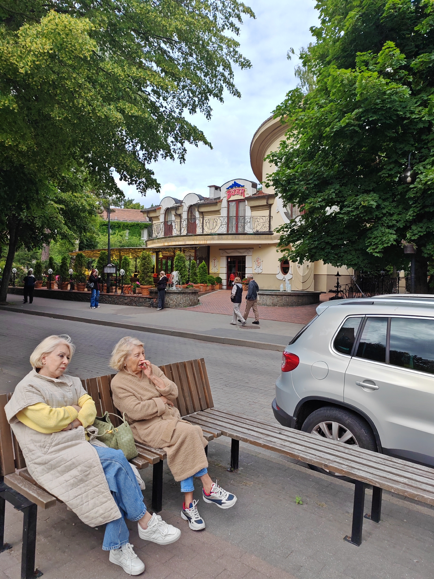 Two elderly women are sitting on a wooden bench on a sidewalk in Svetlogorsk, Russia. The woman on the left is wearing a long, light-colored quilted coat over a yellow sweater and blue jeans, with white sneakers. She has blonde hair and is looking down. To her right, the second woman is wearing a light brown, fluffy coat and blue jeans, with sneakers. She has blonde hair and her hand is near her mouth. A green bag sits between them on the bench.
Behind the bench, a silver SUV is parked next to the sidewalk. Across the street, a light yellow building with a curved facade and arched windows is visible, with a sign that reads "BALTIKA" in red letters. The building appears to be a restaurant or cafe, with outdoor seating areas sheltered by awnings and decorated with potted plants and trees. A small fountain is in front of the building. The scene is set under a cloudy sky, with lush green trees providing shade. Several other people are walking on the sidewalk in the background.