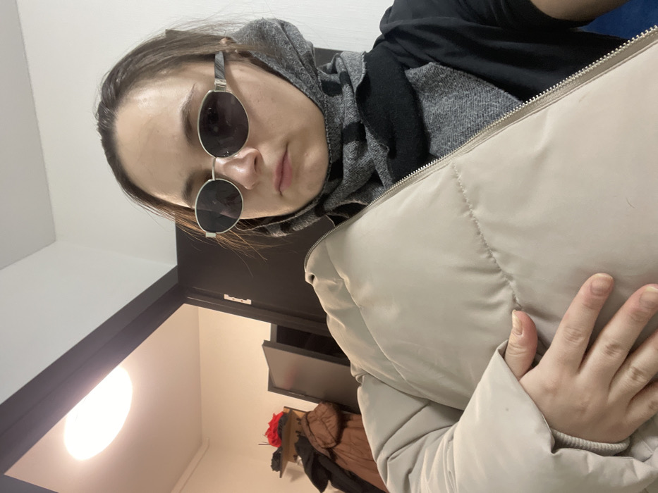 An adult female is captured in a selfie-style, close-up shot, positioned off-center to the right. She wears dark oval sunglasses with silver frames, a black long-sleeved shirt, and a multi-tone grey and black patterned scarf wrapped around her neck and lower face. Her right hand firmly grips a light beige puffer jacket or vest, obscuring part of her left side. The background depicts an indoor residential or commercial environment with white walls. A dark-framed doorway is visible on the left, leading into an adjacent room where a brown coat hangs on a rack, a red object is present, and a dark piece of furniture or open door is seen. An illuminated oval-shaped ceiling light fixture is mounted above the doorway. This scene is situated indoors, within a building in Соболиха, Russia.