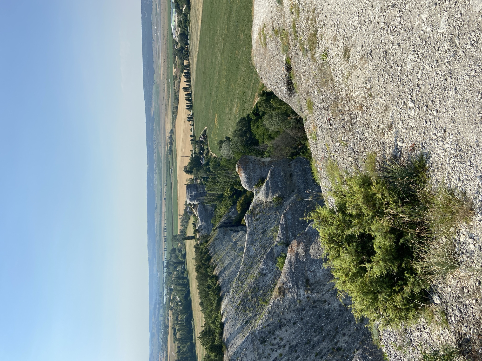 The image depicts a scenic view from a rocky vantage point overlooking a landscape. The foreground is dominated by textured grey rock, with patches of dry grass and a dense green shrub growing out of the rocky surface. The rock formation appears to be a cliff face or a steep incline.
Below the rocky edge, the scene transitions to a terraced landscape with fields of varying colors. Directly below the rocky outcrop, there are dense green trees, forming a verdant band. Further out, there are fields of golden-brown stubble, suggesting recently harvested crops, and vibrant green fields, likely growing crops. A line of trees runs along the edge of one of the fields, creating a distinct visual boundary. A small cluster of buildings is visible nestled within the landscape, suggesting a rural settlement. In the distance, the landscape stretches out, with faint outlines of hills or mountains under a clear, pale blue sky.
The time of day appears to be late morning or early afternoon, given the bright sunlight and the clear sky. The weather seems fair and warm. The overall impression is one of a peaceful, rural setting with dramatic natural rock formations. There is no visible text in the image. The location cues, such as the terraced fields, rural settlement, and rocky terrain, are consistent with a countryside environment.