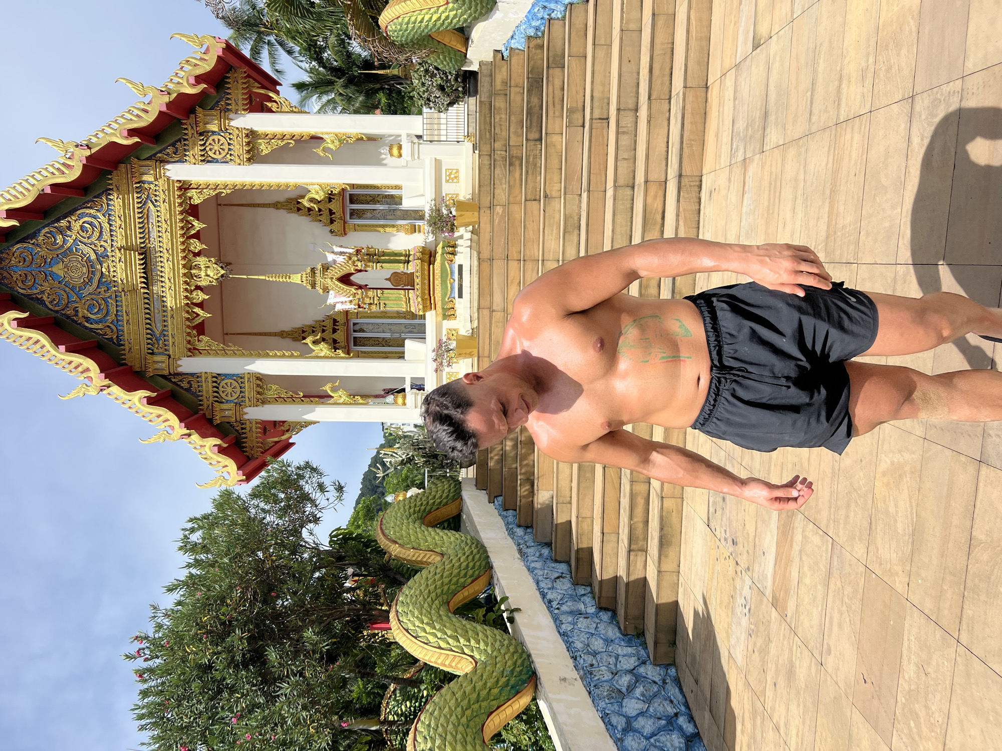 A shirtless man in black shorts stands on a sunlit, tiled walkway in front of a temple in Karon, Thailand. The temple features ornate gold and red trim, with intricate blue and gold detailing on its facade. A large, green serpent statue coils down the side of the temple steps. The man has a tattoo on his chest. The sun casts strong shadows, indicating it is likely midday. The man appears to be looking down, with his head tilted. The background shows lush green trees and a clear blue sky. The overall impression is one of a bright, warm day at a culturally significant site.