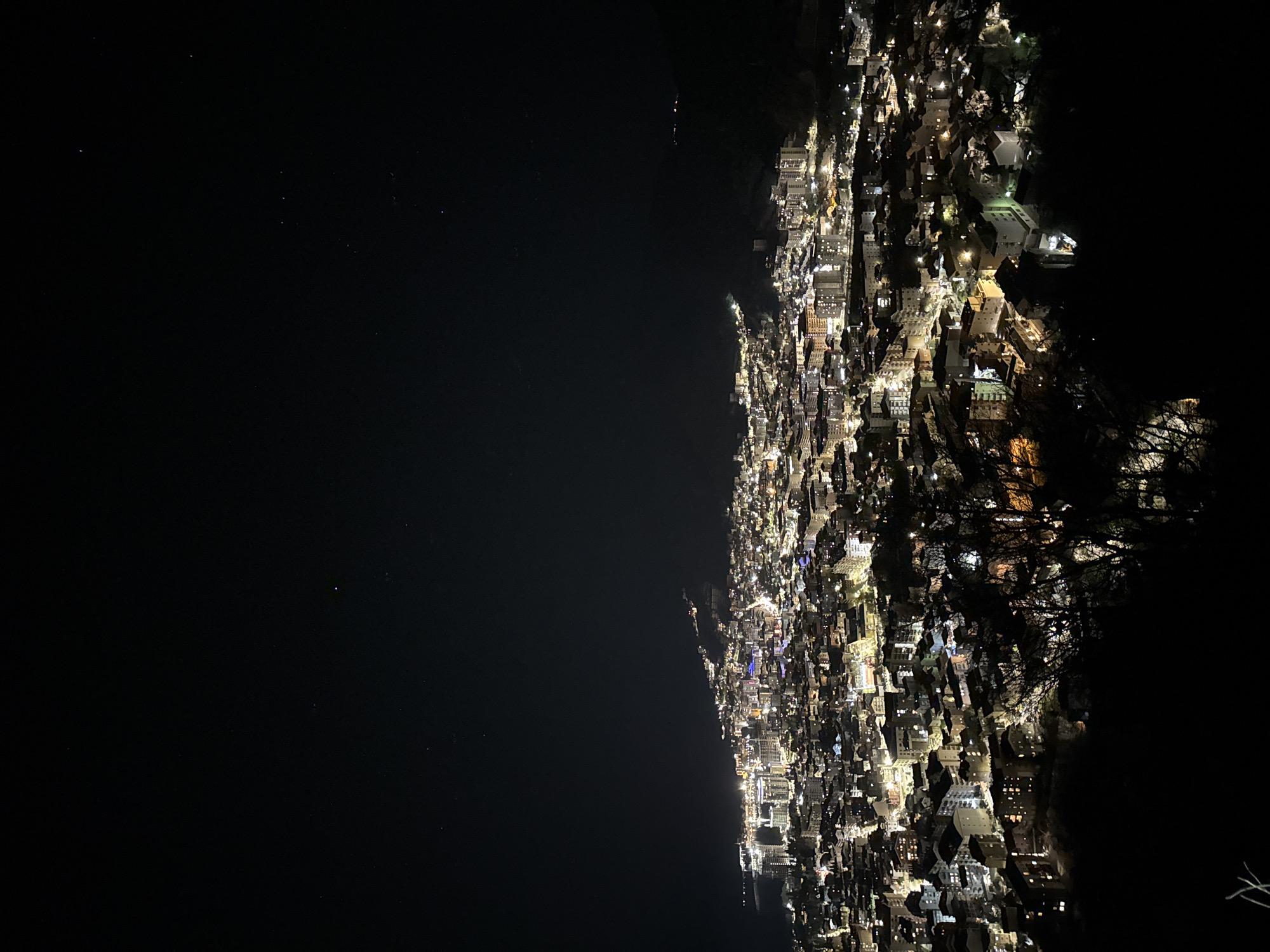 The image is a long-exposure photograph of Budva, Montenegro, at night. The city lights create a bright band along the coastline, contrasting with the dark Adriatic Sea on the left and the dark, obscured mountainsides on the right. Buildings are densely packed, with varied heights and light colors. Scattered brighter points of light suggest specific landmarks or gathering points. The scene is quiet and still, with no visible movement of people or vehicles, conveying a sense of isolation and tranquility.