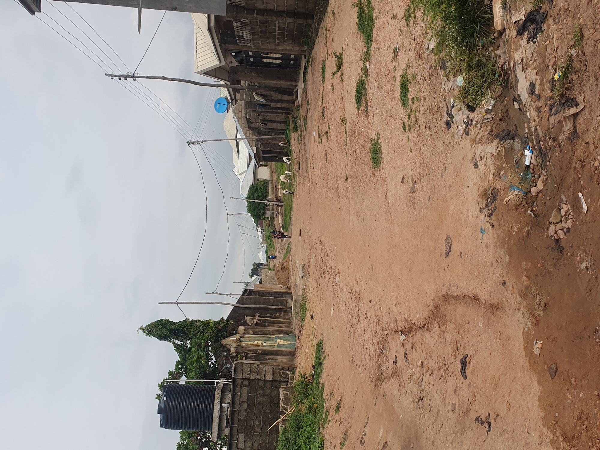 This image depicts a scene in Lokoja, Nigeria, characterized by a dusty, unpaved road. The road is flanked by residential structures made of brick and concrete, with corrugated metal roofs. Vegetation, including trees and shrubs, is present, some growing wild along the roadside. Several goats are visible grazing on the side of the road. In the distance, people are walking along the road. Overhead, power lines and a satellite dish are discernible. Puddles of water and scattered debris, including plastic bags and bottles, suggest a less-than-ideal sanitation situation. A large black water tank is mounted on top of a brick wall on the left side of the image. The overall impression is that of a residential area in a developing urban environment.