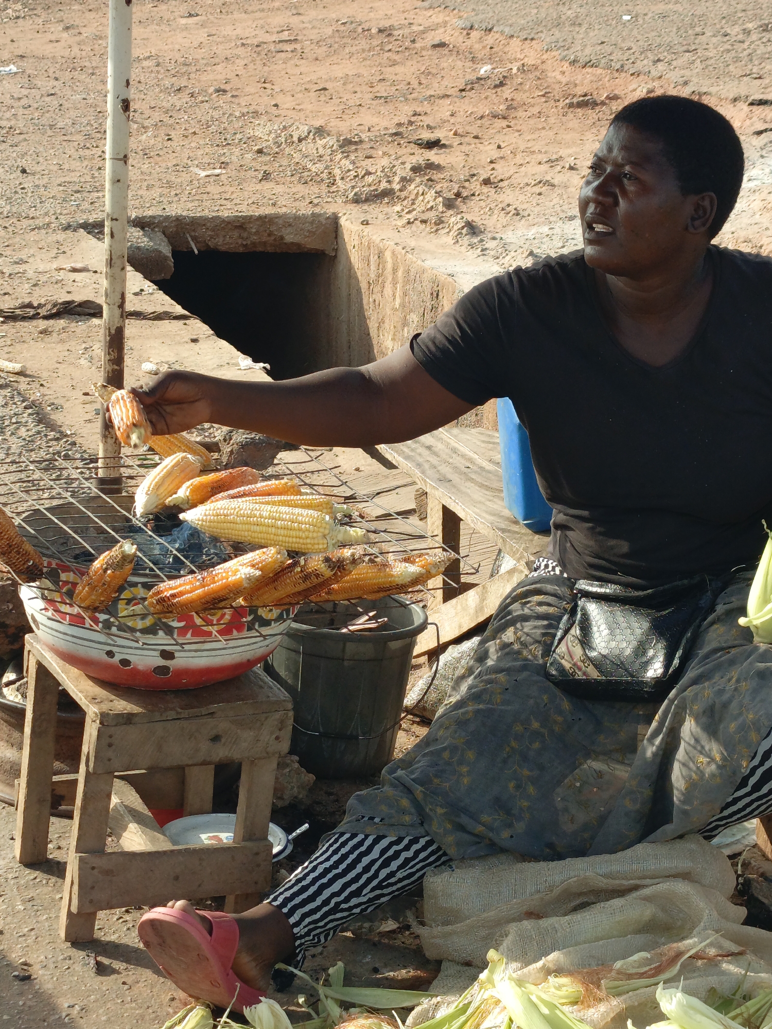 An adult Black woman sits outdoors on a wooden bench in Akwanga, Nigeria. She is wearing a black t-shirt, patterned grey skirt, and black and white striped leggings. A black purse is strapped around her waist. She is offering an ear of corn from a grill on top of a stool next to her. The grill contains multiple ears of corn. A large bowl sits under the grill, and a plastic bucket is next to the stool. Corn husks are scattered on the ground. A metal pole stands to the left of the corn, and there's an open pit in the ground behind it. The ground is dirt.