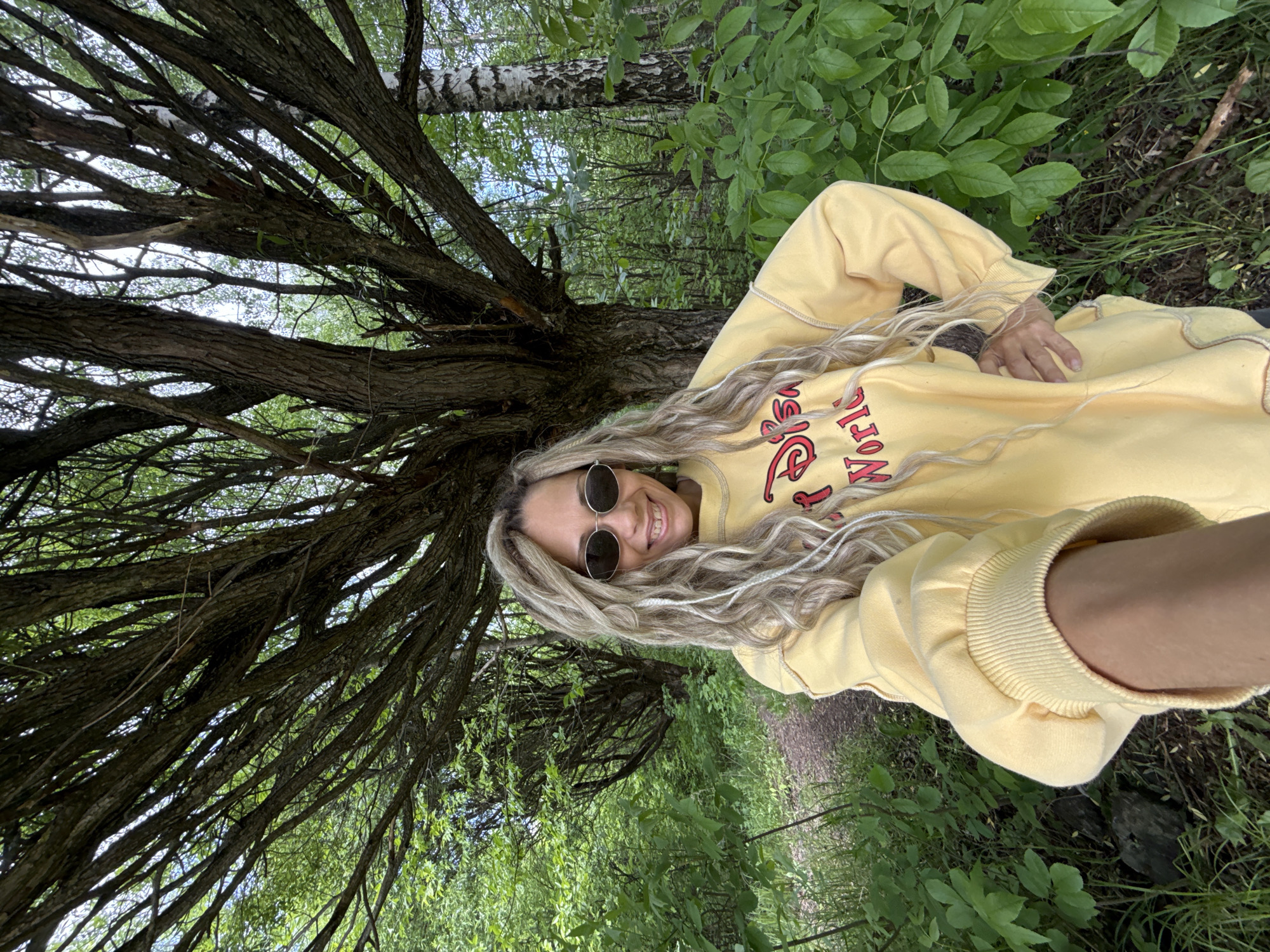 A female with dreadlocks wearing round sunglasses and a yellow sweater is pictured in a forest-like area. She's lying on the ground next to a tree with bare branches that extend across the left side of the frame. There's greenery surrounding her, and a hint of a path can be seen in the background. The location and city are not identifiable from the image.