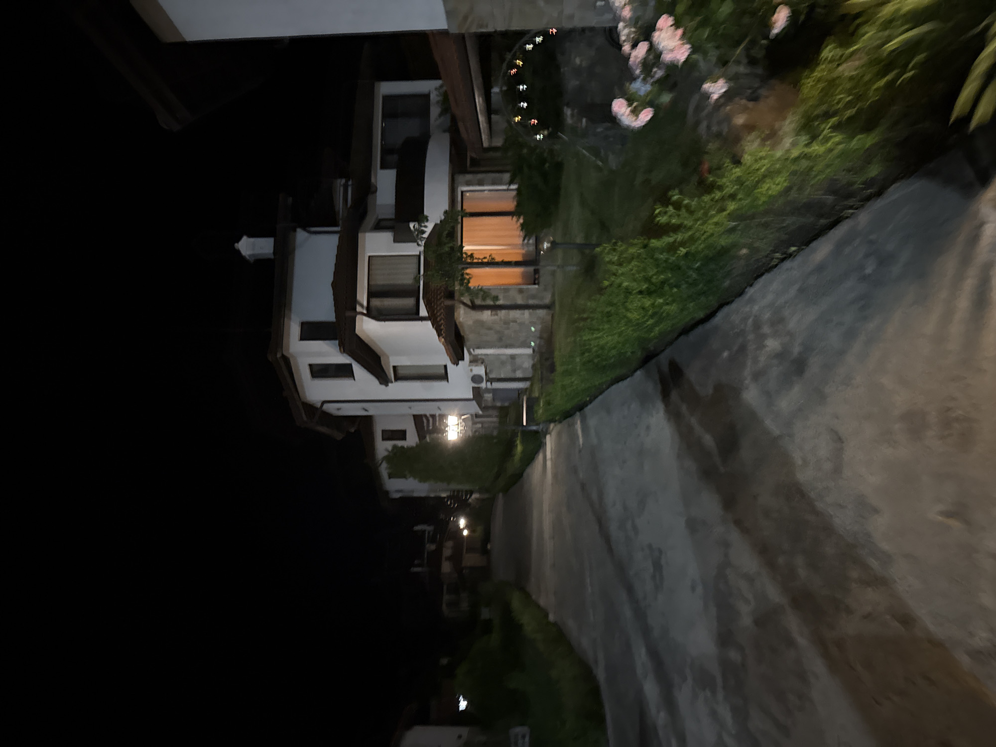 This image depicts a nighttime scene in a residential area within the city of Kosharitsa, Bulgaria. A dark, concrete or paved road ascends gently towards the left, flanked by houses and greenery. On the right side, a prominent multi-story house with white facades and brown tiled roofs features windows, a balcony, and stone cladding on its lower section. Warm light emanates from a ground-floor window of this house. An outdoor air conditioning unit is visible on its left side. In the garden area adjacent to this house, a decorative lamppost illuminates the foreground, revealing green hedges, grass, and pink blooming flowers. Multi-colored string lights are strung over an archway or structure further into the garden. On the left side of the road, more white-walled houses with brown roofs are visible further up the incline, one featuring a white chimney. The dark sky is clear. No individuals or active subjects are present in the scene.