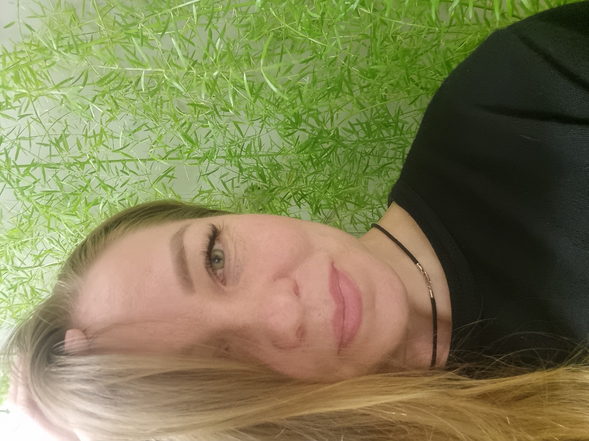 A close-up, diagonal portrait features a woman with light blonde hair and light-colored eyes, set against a vibrant green foliage backdrop. The woman is positioned with her head tilted, and her blonde hair cascades towards the bottom of the frame. She has a subtle, pleasant smile and looks directly towards the viewer. Her skin appears fair, and she wears a black top or sweater, complemented by a thin black choker necklace with a small silver clasp visible on her neck. The background consists entirely of dense, delicate green leaves, possibly from an asparagus fern or similar plant, creating a natural and soft setting. The lighting is soft and even, suggesting an indoor environment or diffused outdoor light. There is no visible text in the image. The scene is static, capturing a posed moment in Kemul, Russia.