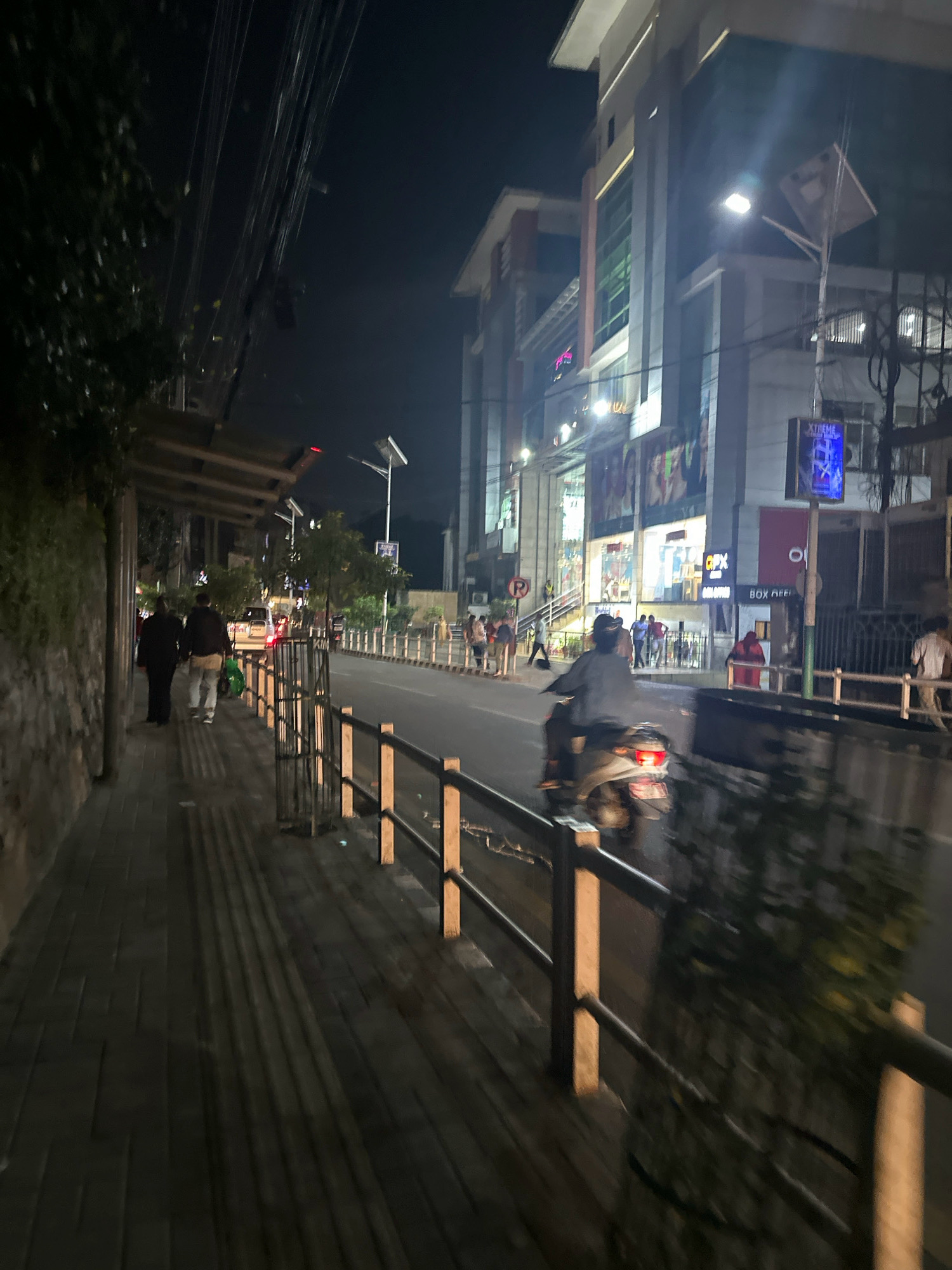 A street scene unfolds at night in Kathmandu Metropolitan City, Nepal. The perspective is from a sidewalk, looking across a moderately busy street towards a multi-story commercial building.
On the sidewalk to the left, two individuals are walking away from the camera, their figures silhouetted against the ambient light. The sidewalk is paved with dark rectangular tiles, casting long shadows from an unseen light source. A stone wall borders the sidewalk on the far left.
The street itself is illuminated by streetlights and the lights emanating from the building. Traffic appears to be light, with a scooter visible in the mid-ground, its rider moving away from the camera. Several other vehicles, including a white car, are parked or moving in the distance.
The building on the right is brightly lit, showcasing advertisements and storefronts. A prominent sign with the word "XTREME" is visible. Fencing and railings line the edge of the sidewalk separating it from the street. Trees and foliage are present along the street, adding greenery to the urban landscape. The overall impression is of an active commercial area after dark.