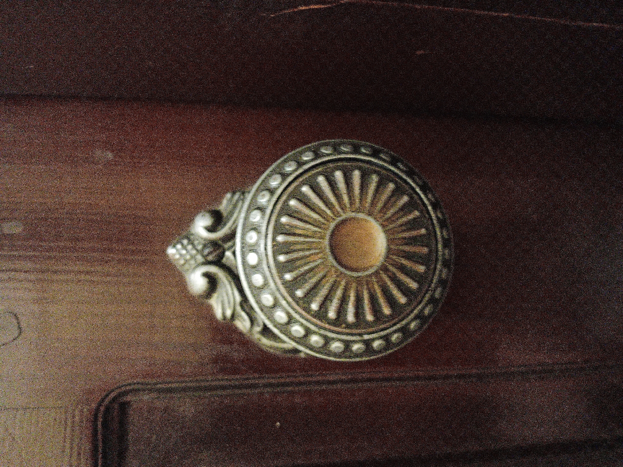 The image is a close-up, high-angle shot of an ornate, antique door knocker mounted on a dark wooden door. The knocker is made of metal, likely brass or bronze, and features a detailed, circular design with radiating spokes and decorative elements at the top. The wooden door appears to have a visible grain and is a dark, rich brown color. The lighting is somewhat dim, creating shadows that highlight the texture of both the door and the knocker. The overall impression is one of historical significance and traditional craftsmanship. There is no visible indication of the city of Lutsk, Ukraine, within the image itself.