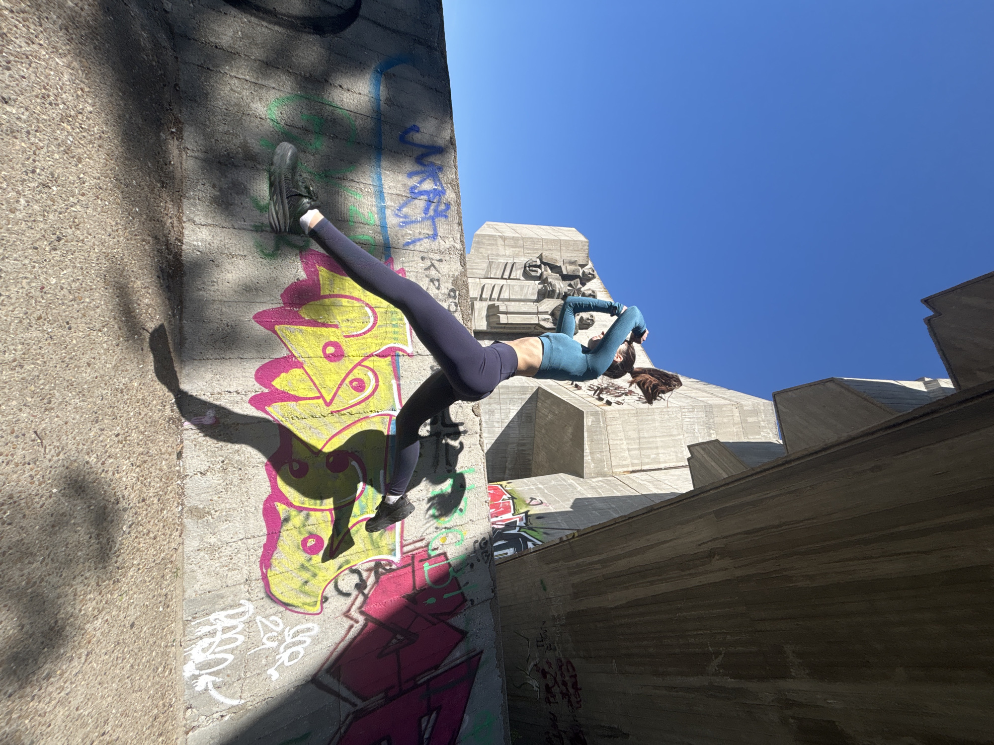 This dynamic, low-angle photograph captures a person performing an athletic pose against a heavily graffitied concrete wall in an urban environment. The subject, likely a female, is dressed in dark leggings, a teal long-sleeved crop top, and black athletic shoes with white socks. She is positioned horizontally, appearing to be suspended or leaning against the wall, with one foot pressed against the concrete and the other leg extended. Her torso is slightly arched, and her arms are bent with hands near her head, suggesting a flexible or strong hold. Her long, dark hair is visible towards the right side of the frame.
The setting is characterized by monumental, Brutalist-style concrete architecture, with massive, angular structures rising towards a clear, bright blue sky. The wall the person is against, as well as other concrete surfaces, is extensively covered in vibrant graffiti, featuring stylized lettering and designs in yellow, pink, green, and blue, providing a colorful contrast to the stark gray concrete. Strong, directional sunlight casts prominent shadows across the textured surfaces and the person, indicating daytime, likely midday or early afternoon, with excellent weather.
The image evokes a sense of urban exploration, agility, and artistic expression, blending human movement with the raw aesthetics of concrete and street art. The only discernible text is the brand name "NIKE" faintly visible on the sole of the subject's shoe.