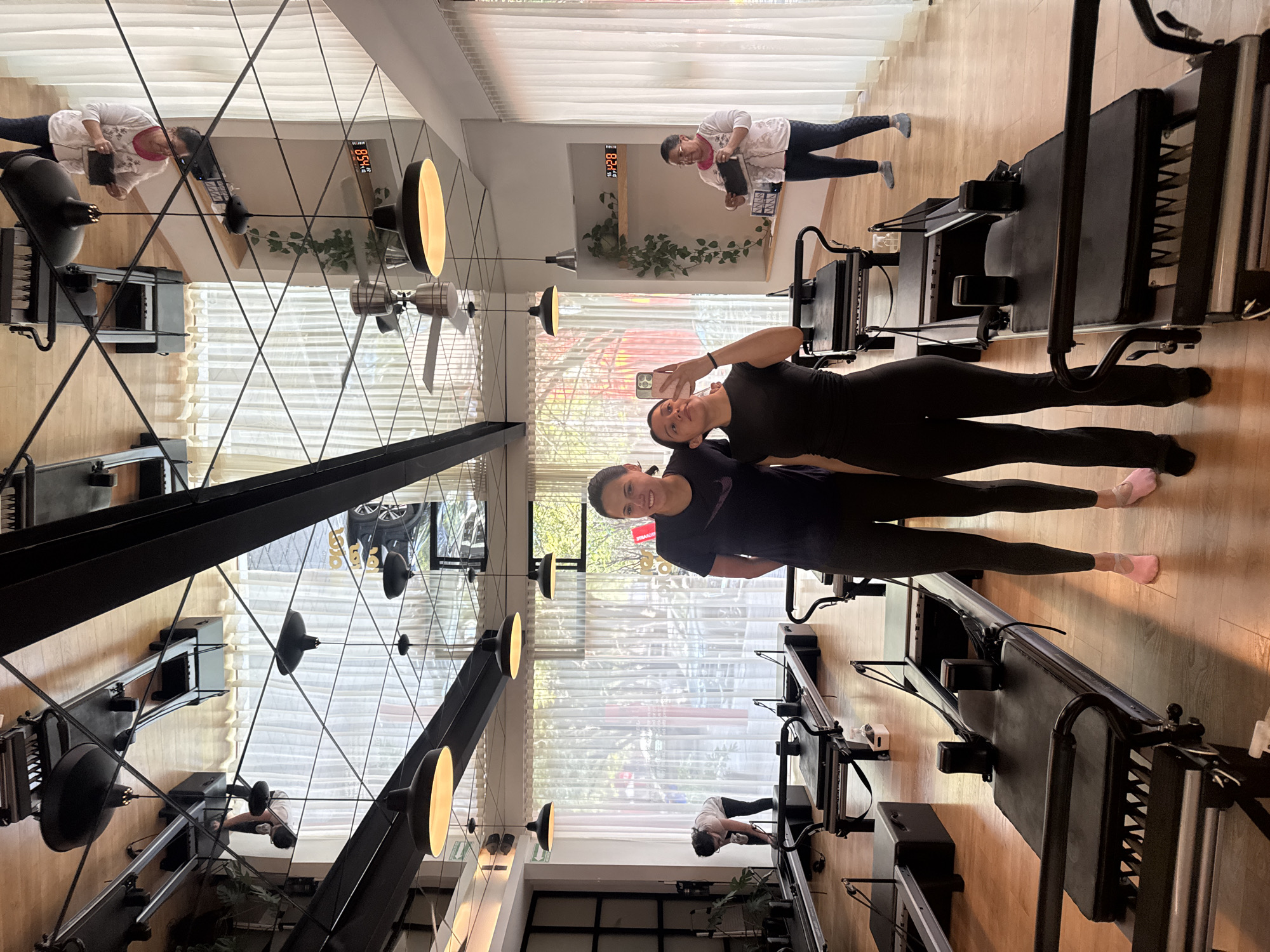 Two young women pose for a selfie in a Pilates studio in Puebla, Mexico. The studio is equipped with several Pilates reformer machines. An older woman stands on one of the machines in the background, holding a tablet. The studio has a large window with white curtains and a mirrored wall. An analog digital clock displays "15:54" on the wall.