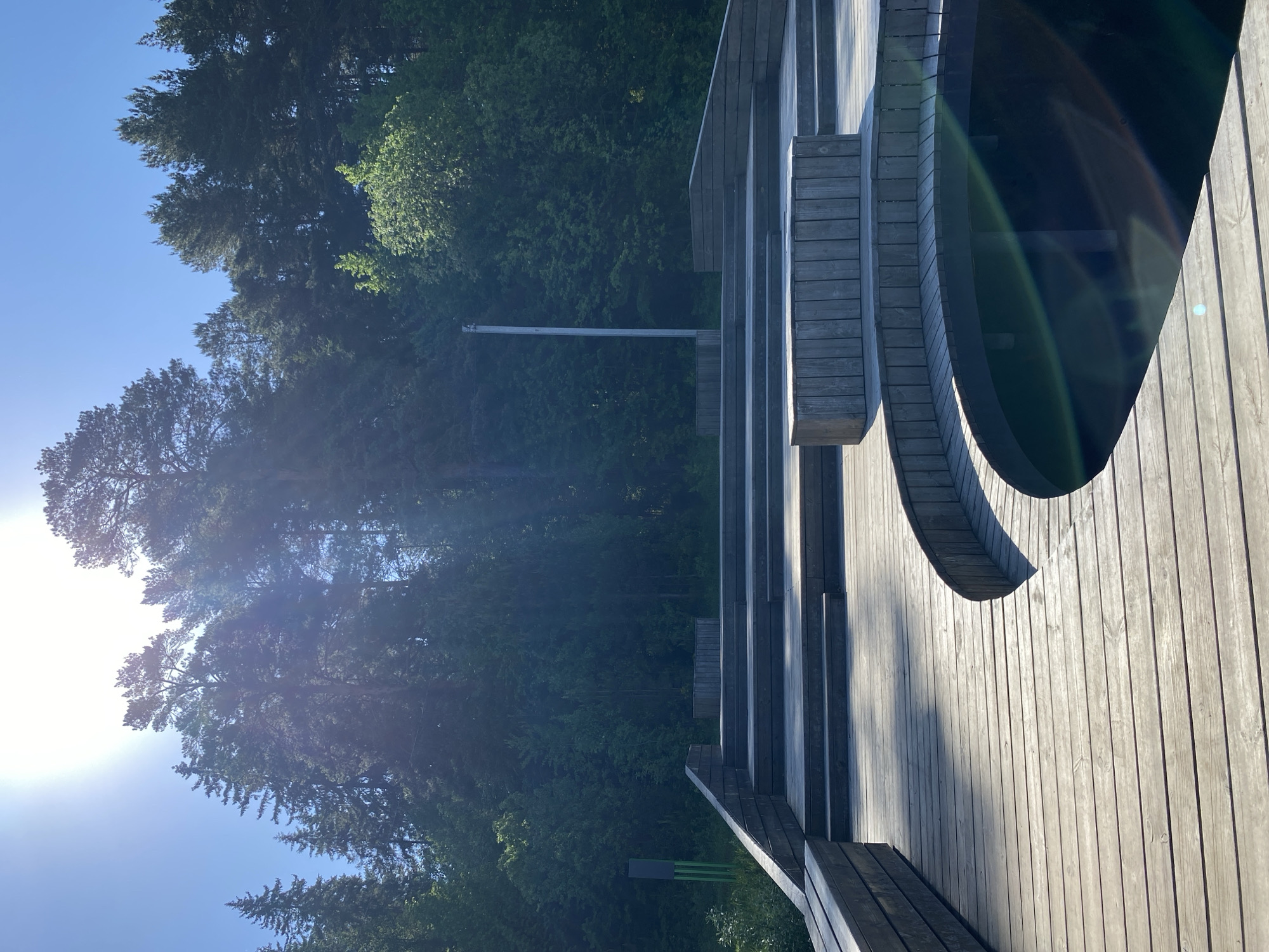 The image depicts a wooden structure, possibly a viewing platform or deck, constructed from light-colored wood planks. The structure features curved sections and built-in benches. In the background, there is a forest with a mix of green foliage and tall trees, under a clear blue sky. The sun is visible in the lower left corner of the image, creating a lens flare effect.
There is no direct evidence in the image to confirm its location in Votkinsk, Russia. However, the presence of a wooded area adjacent to a wooden structure is consistent with park areas or recreational spaces that might be found in various locations.