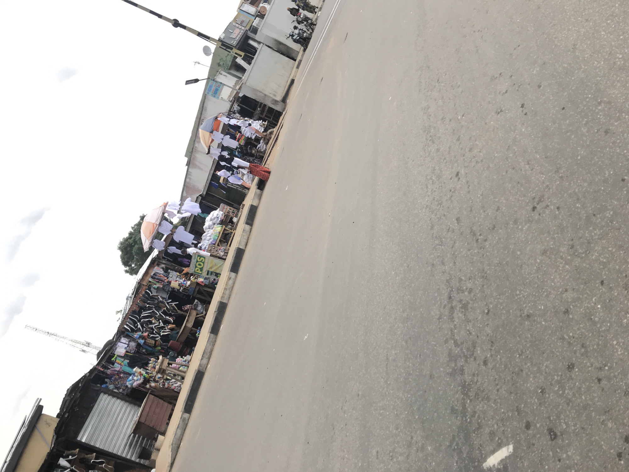 The image depicts a bustling street market scene in Mandate Estate, Ilorin, Nigeria. A wide, grey asphalt road occupies the right side and foreground, showing some texture and faint markings, including a white line at the bottom right. This road is bordered on the left by a black and white striped curb, separating it from the active market area.
Along the left side of the street, numerous makeshift market stalls are visible. These structures are constructed from wood and corrugated iron sheets, creating shaded areas for vendors and their goods. A wide variety of merchandise is on display, including rows of white shirts or garments hanging neatly, some under large umbrellas (one light blue and orange, another white). There's a prominent display of shoes, specifically black and white sneakers, stacked on shelves. Other stalls feature household items, plastic goods, and general merchandise, indicating a diverse array of products available for sale.
Several individuals are present within the market, appearing to be both vendors and shoppers, engaging in daily commercial activities. Some are standing under the umbrellas, while others move between the stalls. Further down the street, two motorcycles are parked on the side, with a few people standing nearby.
The background reveals concrete buildings, some appearing weathered, suggesting established businesses or residences behind the market. The sky is overcast with grey clouds, indicating a cloudy daytime setting. A tall metal structure, possibly a telecommunications mast or a construction crane, is visible in the far-left background. Streetlights and utility poles are also part of the urban infrastructure.
Visible text includes a distinct green sign with "POS" (Point of Sale) written in white letters, affixed to one of the stalls. Another partially obscured blue sign appears to have text resembling "ORIENT." The overall scene captures the vibrant atmosphere of an active local market.