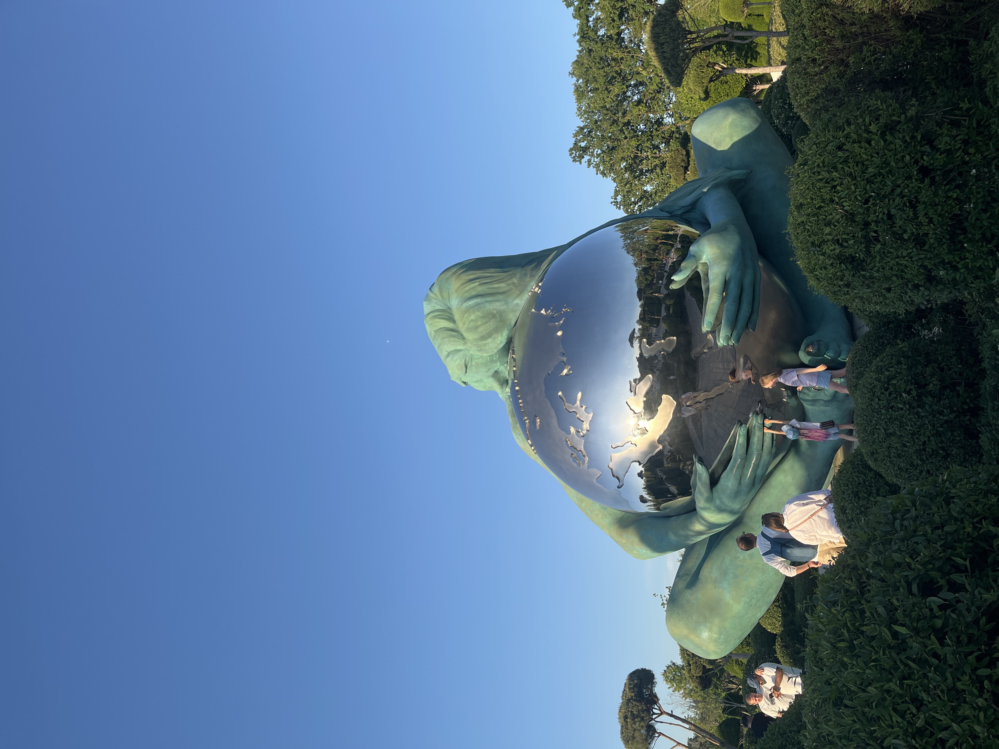 This media file captures a large, teal-colored statue of a reclining figure, resembling a woman or mythical entity, gracefully holding a massive, reflective silver globe. The globe is intricately etched with landmasses, depicting the Earth. The statue is situated outdoors in a well-maintained park or garden in Krasnodar, Russia, surrounded by lush green hedges and trees with manicured foliage.
Several people, including adults and children, are gathered around the base of the statue. Children are seen reaching towards the shimmering globe, while adults observe the artwork or take photographs. One person in a white shirt is bending down near the statue's base. The scene takes place during a clear, sunny day, with a vibrant blue sky visible above. Strong sunlight creates distinct reflections on the globe's surface, showing inverted images of the surrounding trees, sky, and people. The overall atmosphere suggests visitors are admiring the unique sculpture in a pleasant outdoor setting. There is no visible text in the image.