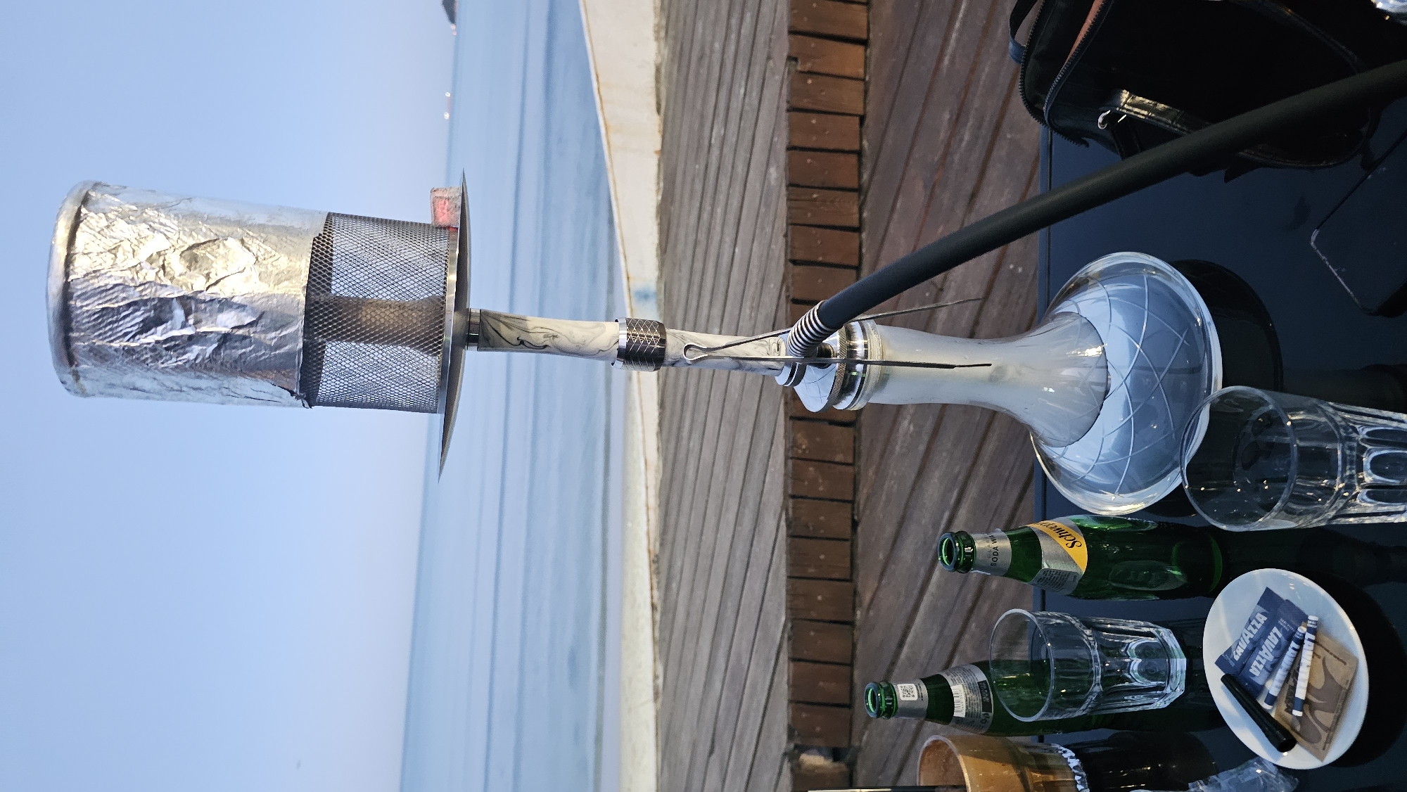 The image shows a hookah on a black table. There are two green Schweppes bottles, two patterned glasses, and a small white plate with Lavazza sugar packets and stirrers on the table. The base of the hookah is translucent, and the hose is black. The bowl of the hookah is covered with foil and a metal screen.
In the background, there is a wooden boardwalk and the sea. The location is likely near the coast in Varna, Bulgaria.