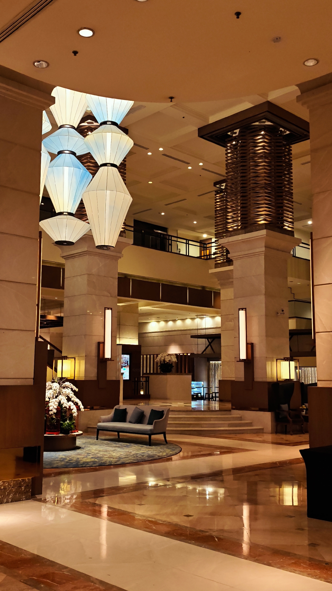 This media file depicts the interior of a luxurious hotel lobby in Hanoi, Vietnam. The scene is bathed in warm, ambient light, suggesting it's either evening or the lighting is designed to create a cozy atmosphere.
The central focus is the grand architecture of the lobby. Massive marble pillars, intricately carved wooden accents on a tall structure, and a high ceiling adorned with recessed lighting create a sense of opulence. Dominating the upper part of the frame are large, stacked lantern-like light fixtures, some glowing white and others with a subtle blue hue, adding an artistic and dramatic element to the space.
In the mid-ground, a comfortable-looking grey sofa with dark pillows sits on a circular, patterned rug, suggesting a seating area for guests. Beside it, a large floral arrangement with white orchids and red accents adds a touch of natural beauty and color. The floor is a polished marble with a reflective surface, showing the warm glow of the lights and the architectural details. Stairs are visible in the background, leading to another level of the hotel.
The overall impression is one of elegance, sophistication, and tranquility. The lack of visible people suggests a quiet moment in the lobby, perhaps early morning or late at night. There is no visible text in the image. The scene conveys a sense of luxury and comfort, characteristic of a high-end hotel. The interior design elements, particularly the lighting and materials, hint at a fusion of modern aesthetics with traditional influences that might be found in Vietnamese hospitality.