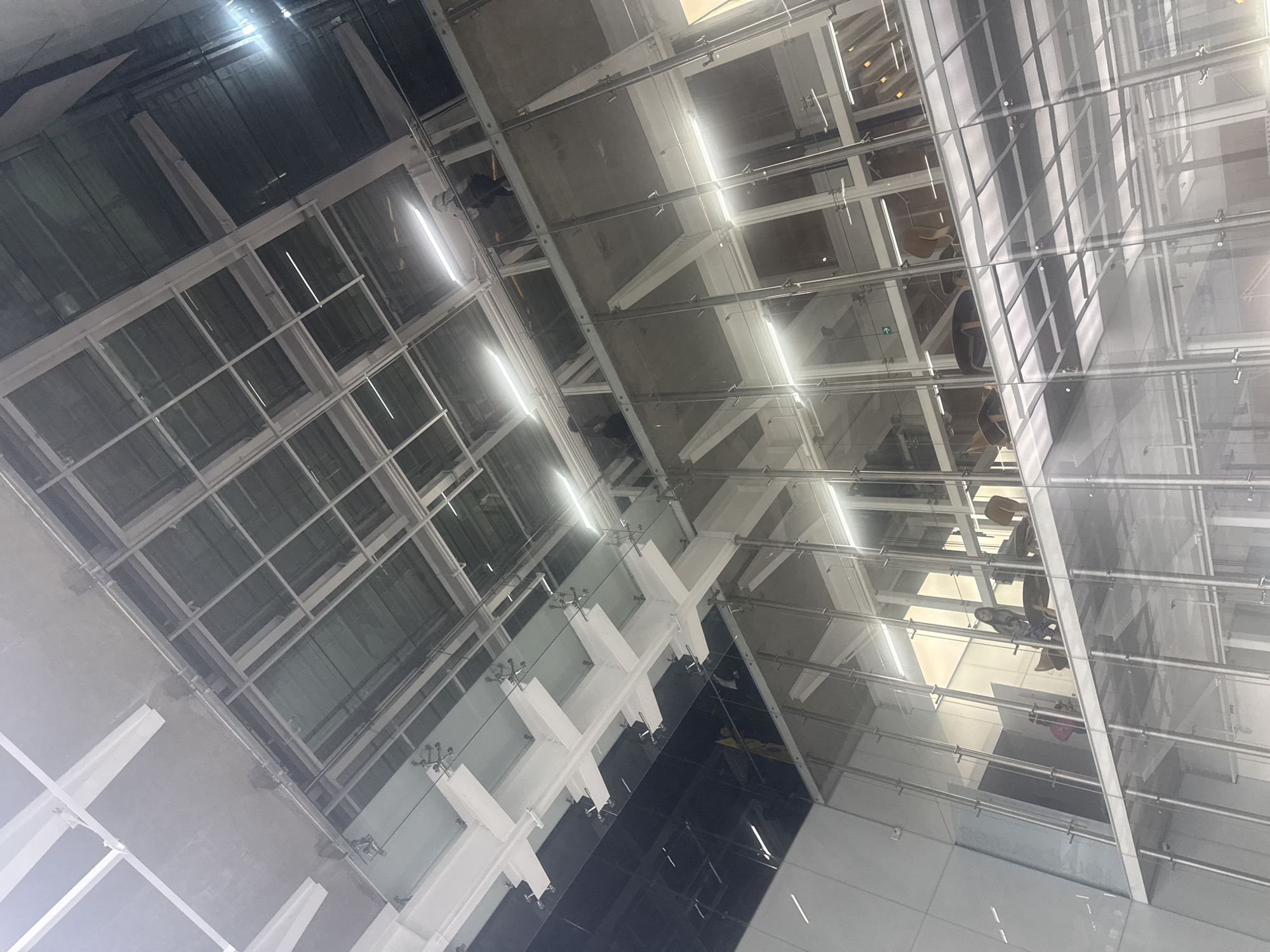 The image shows an interior view of a building, likely taken from a lower level looking upwards. The architecture features a combination of glass and steel construction, with a visible grid-like structure and supporting beams. Reflections in the glass surfaces provide glimpses of people and furniture on upper floors. The lighting appears to be a mix of natural and artificial, with bright highlights. The location is presumed to be in Puebla, Mexico.