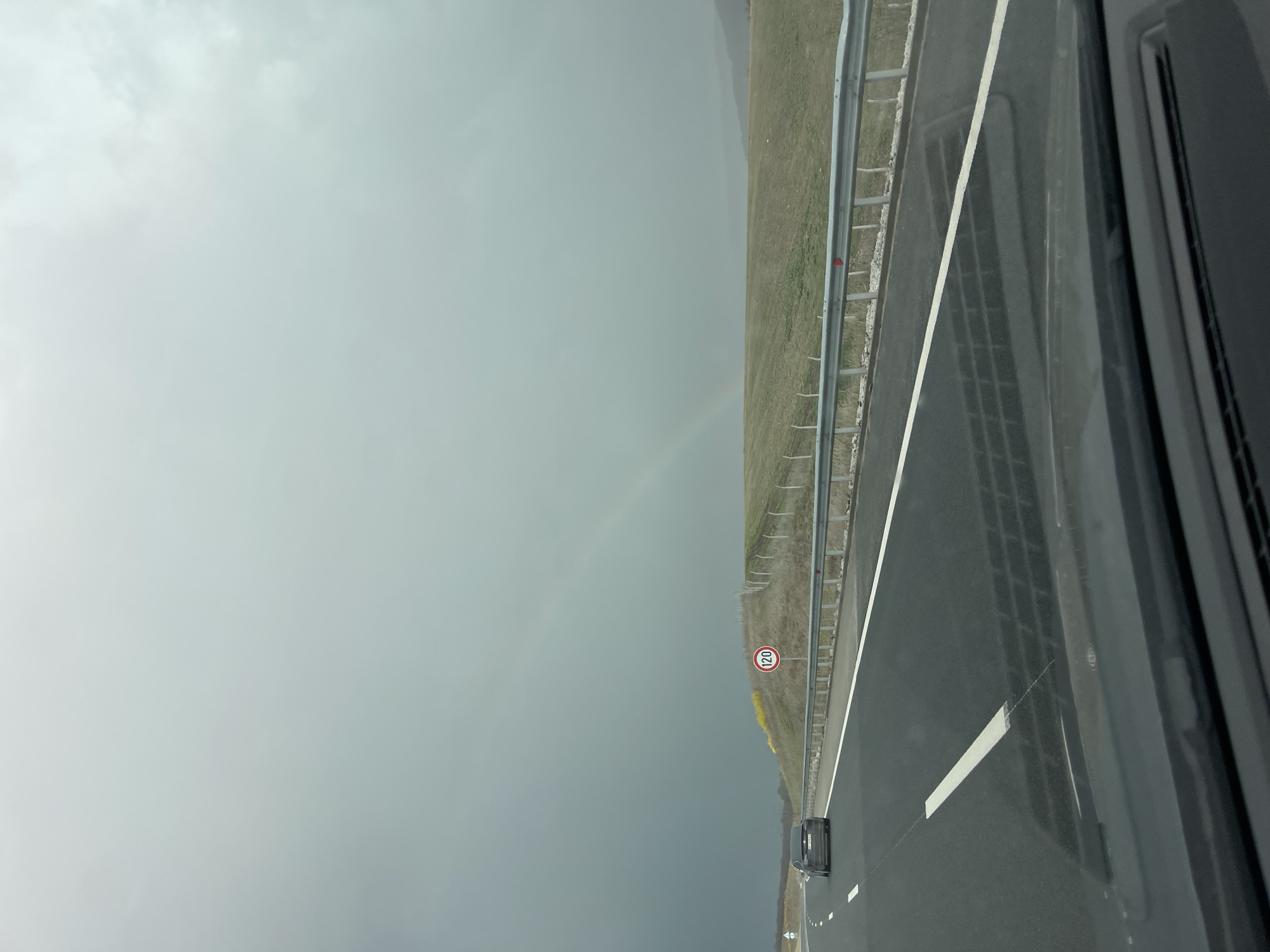 The image shows a highway in Buhovtsi, Bulgaria, possibly taken from inside a moving vehicle. A gray sedan is seen traveling on the road. A metal guardrail runs along the right side of the highway with a speed limit sign of 120. In the distance, a faint rainbow is visible in the sky above the adjacent field. The weather appears overcast.