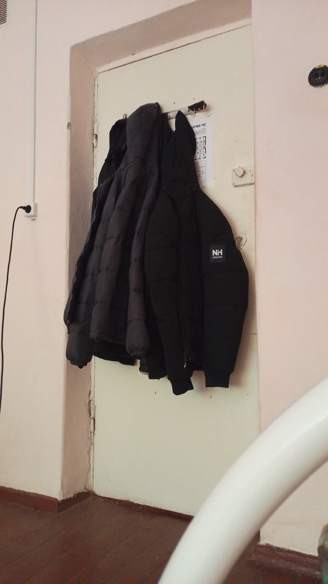 The image shows several dark-colored jackets hanging on hooks attached to a white door. The jackets appear to be puffer jackets, with one prominently displaying a white "NH" logo. The door is situated in a hallway with light-colored walls and a wooden floor. To the left of the door, a black electrical cord is plugged into a white wall outlet. A white, curved object, possibly a railing or furniture, is visible in the foreground, partially obscuring the lower right portion of the scene. The overall impression is of an interior space, likely a hallway or entrance, within a building in Tomsk, Russia.