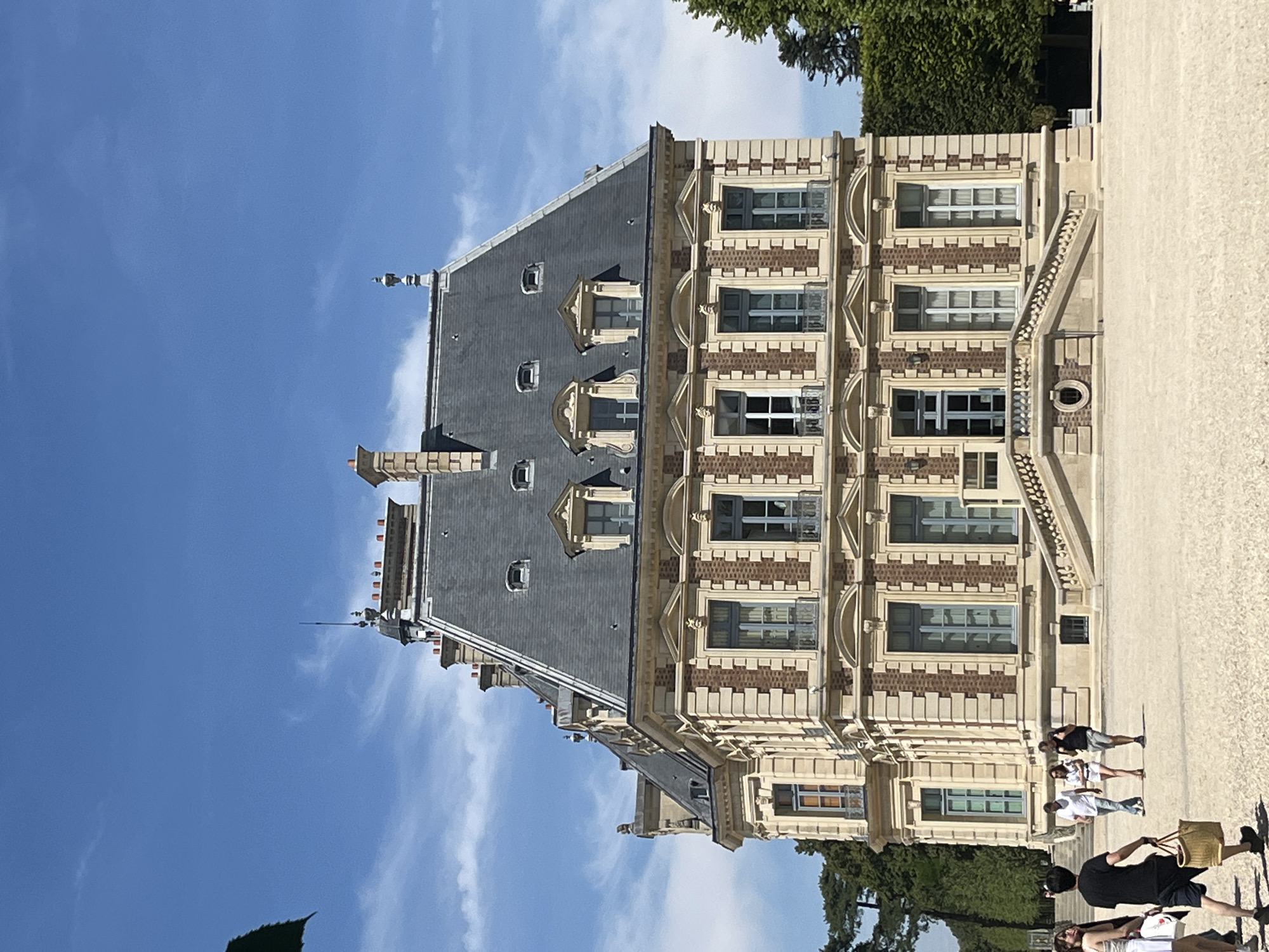 The image shows a large, ornate building, identified as the Pavillon de l'Horloge at the Château de Sceaux, located in the city of Sceaux, France. The building features a mansard roof covered in dark slate tiles, with dormer windows on the upper levels. The facade is constructed with alternating bands of light-colored stone and reddish-brown brick, accented by decorative stonework around the windows and cornices. A grand staircase with balustrades leads up to the main entrance of the building.
In the foreground, a few people are visible walking on a gravelly courtyard. One person in the immediate foreground, facing away from the camera, carries a woven basket. Other individuals are further back, appearing to be tourists. The sky is a clear, bright blue with scattered white clouds. To the right of the building, a manicured green hedge and trees are visible, suggesting a park or garden setting. The overall scene depicts a sunny day at a historical French estate.