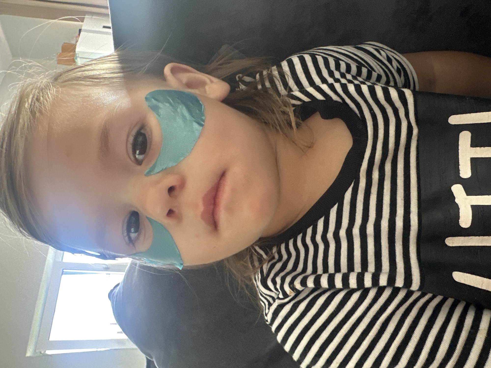 A close-up, eye-level, indoor shot shows a young child with blonde hair and green under-eye patches. The child is wearing a black and white striped t-shirt. The background is dark and out of focus, with a hint of a window and furniture visible. The scene is likely a home environment. The image does not provide any information about the city of Kharnavy, Russia.
