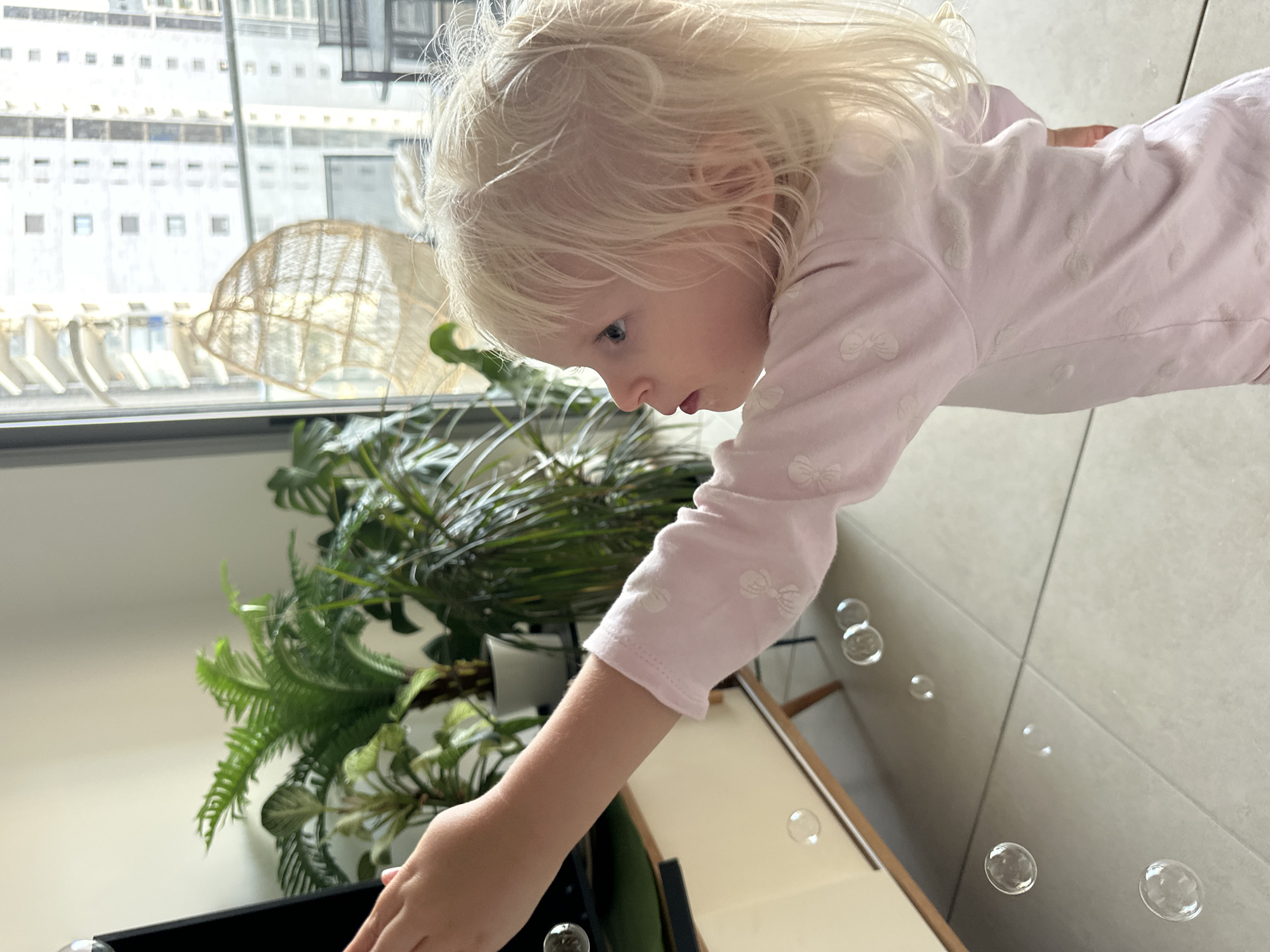A young girl with blonde hair is reaching out her left hand towards bubbles in the air. She is wearing a light pink long-sleeved shirt with a subtle pattern of what appear to be bows. The background shows a large window with a cityscape visible outside, indicating an urban setting. There are several houseplants in pots near the window and a piece of furniture. The floor is tiled. This scene is taking place indoors in the city of Petah Tikva, Israel.