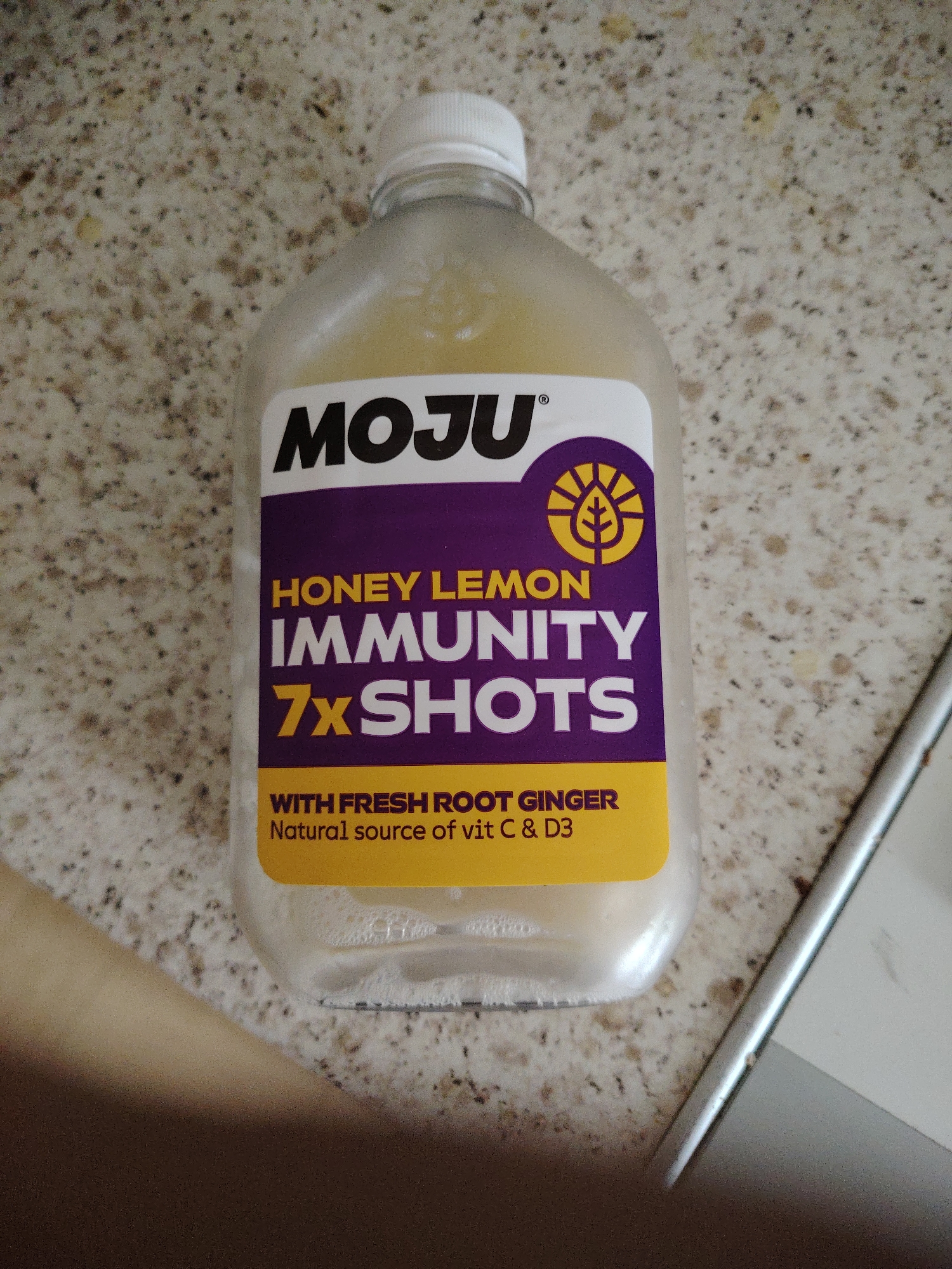 The image depicts a translucent plastic bottle containing a pale yellow, cloudy liquid, situated on a speckled countertop. The requested location, Huntingdonshire, United Kingdom, cannot be confirmed by visual evidence within the image, which solely depicts an indoor setting.
The bottle, centrally positioned and slightly angled to the left, has a white, ridged screw-top cap. The liquid inside has a visible head of foam/bubbles at the top. A prominent label wraps around the bottle. The upper section of the label is dark purple, featuring the brand name "MOJU" in large black capital letters with a registered trademark symbol "®" next to the 'U'. To the right of the brand name, a yellow circular logo with a stylized plant or sunburst design is visible. Below the brand name, white text reads "HONEY LEMON IMMUNITY," with "7x SHOTS" in larger yellow text immediately below it. The bottom section of the label is solid yellow, containing black text that states "WITH FRESH ROOT GINGER" and, in a smaller font, "Natural source of vit C & D3."
The countertop surface is a light, variegated granite or composite material, exhibiting numerous small, irregular flecks of dark brown, black, and lighter tan/beige. The lighting appears to be from above, creating subtle reflections on the bottle and the countertop. To the immediate right of the bottle, a vertical strip of reflective silver-colored metal, possibly the edge of an electronic device like a laptop or tablet, is visible. The lower left portion of the image is blurred, showing a tan or light brown surface, potentially another piece of furniture or flooring. No individuals are present in the frame.