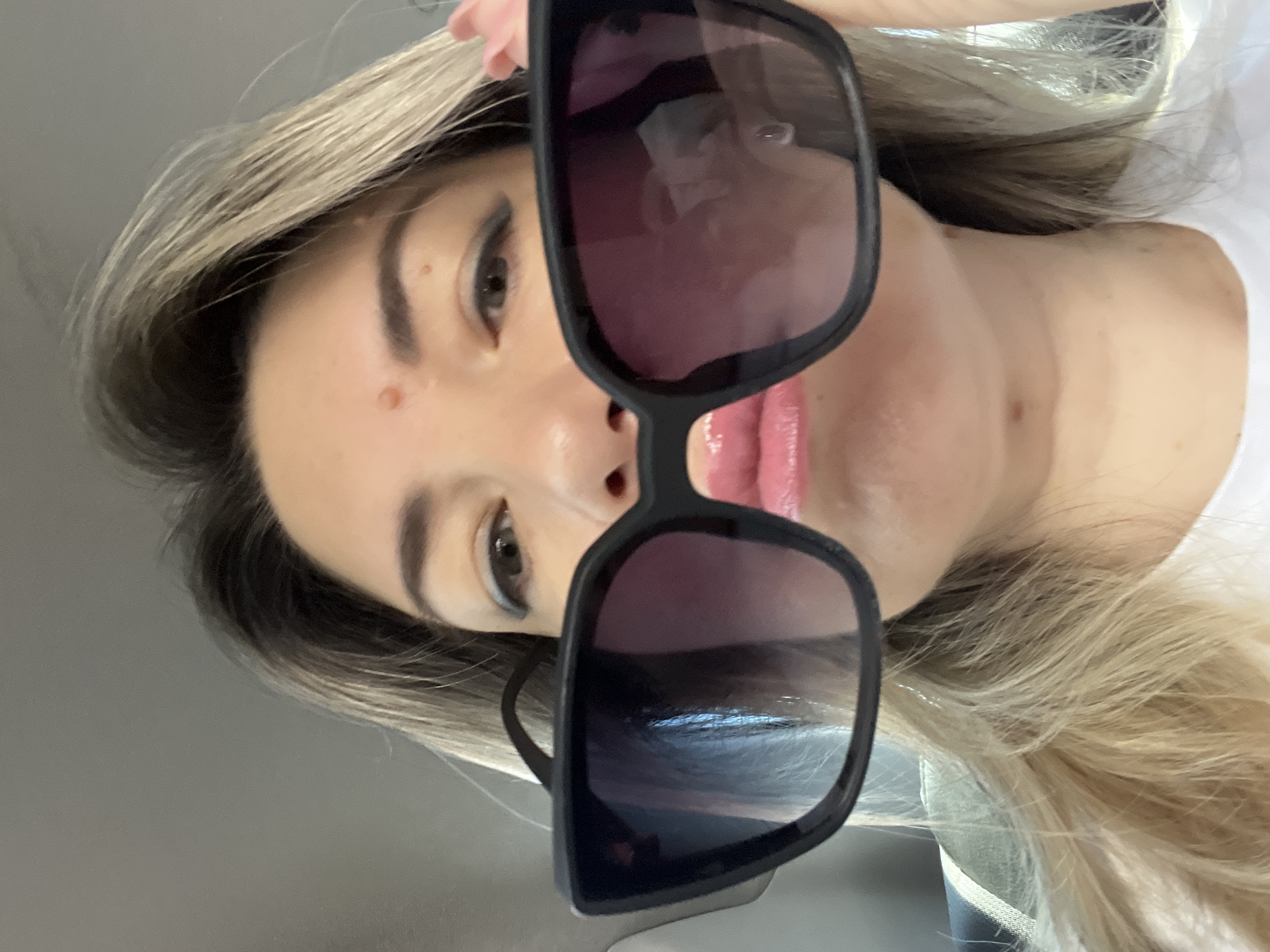 This is a close-up, selfie-style photograph of a young woman in what appears to be a vehicle. She is holding a pair of large, dark-rimmed sunglasses in front of her face, obscuring her nose and part of her mouth. Her eyes are visible, with subtle dark eyeliner. Her lips are slightly pursed. She has fair skin with a mole on her forehead and another on her neck. Her hair is blonde and appears to be styled down. The background suggests the interior of a car, with hints of a headrest and a seatbelt buckle. The lighting is natural, likely from daylight filtering into the car. The specific location in Sochi, Russia is not discernible from the image.