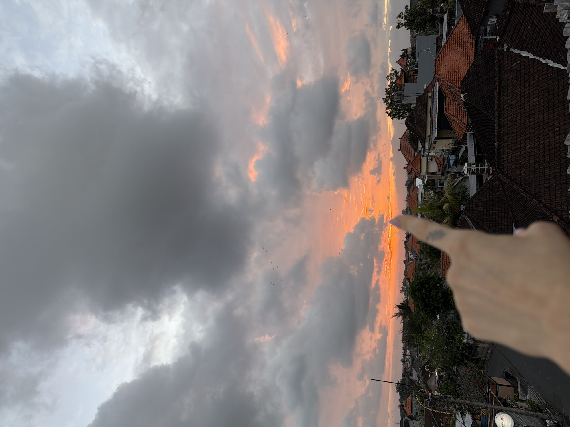 An aerial, over-the-shoulder view shows a finger pointing towards a dramatic sunset over a cityscape. The sky is filled with a mix of dark, heavy clouds and streaks of vibrant orange and pink light, indicating the sun is low on the horizon. The landscape below consists of numerous buildings with terracotta tiled roofs, characteristic of North Kuta, Indonesia. A street is visible between some of the houses. The overall impression is one of a beautiful, yet somewhat overcast, sunset experience.
