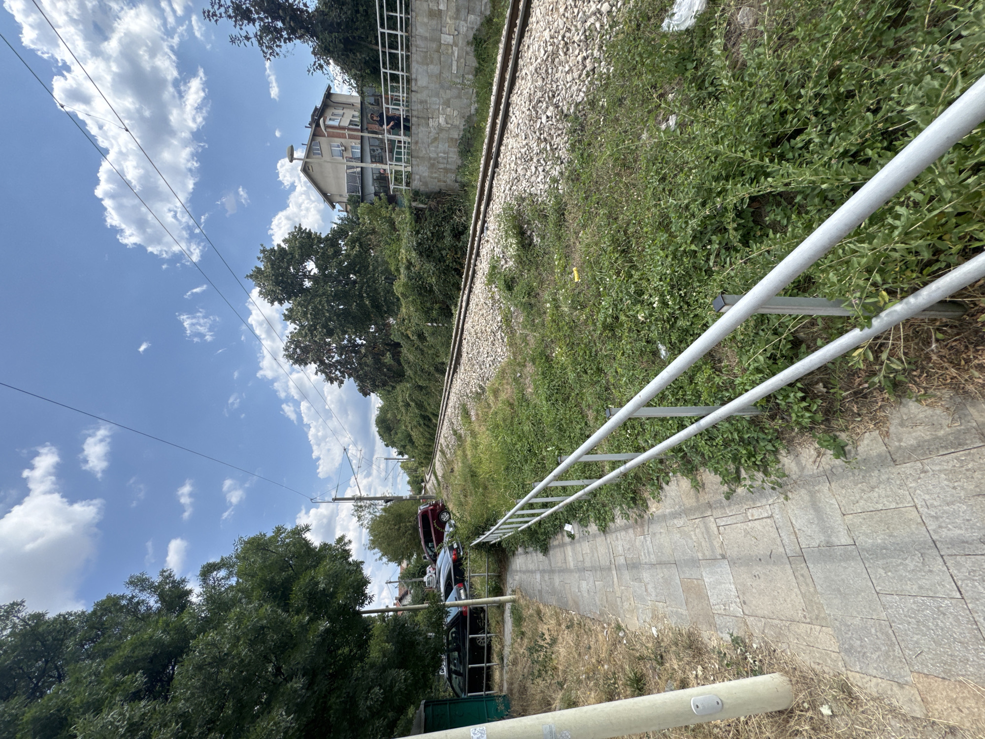 The image shows a pedestrian path alongside a set of train tracks in Sofia, Bulgaria. On the left, a paved walkway runs parallel to a grassy area with some dry vegetation and a white pole. Beyond the grass, several parked cars, including a black sedan and a red SUV, are visible next to a green structure. The train tracks, covered in gravel, curve to the right and are bordered by dense green foliage and trees. In the background, a multi-story building with a light-colored facade and red-brick accents stands on a raised platform with a white railing. Two individuals are standing on this platform, and a street lamp is mounted on the building. The sky above is blue with scattered white clouds.