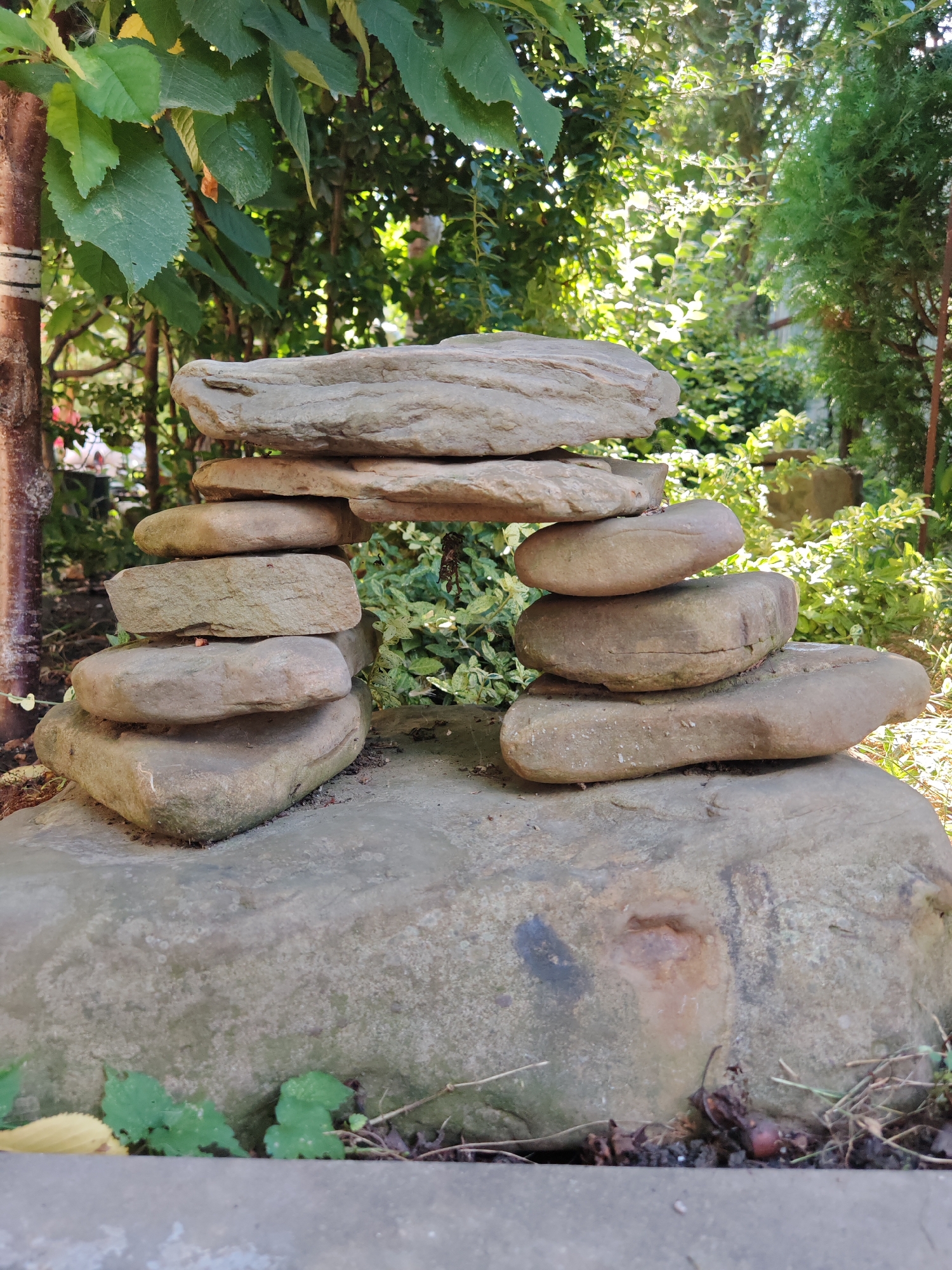 A rock structure, possibly a garden decoration, is the main subject of the image. It is constructed from several flat, irregularly shaped stones stacked on top of each other, creating a somewhat arched form. The structure sits on a larger, flatter rock. The background consists of lush green foliage, trees, and sunlight filtering through the leaves, suggesting an outdoor, possibly garden or park setting. The ground around the rocks is covered with soil and some dry leaves. The image is taken from a slightly low angle, focusing on the rock arrangement. The location of Oktyabrskiy, Russia is not visually discernible from the image itself.