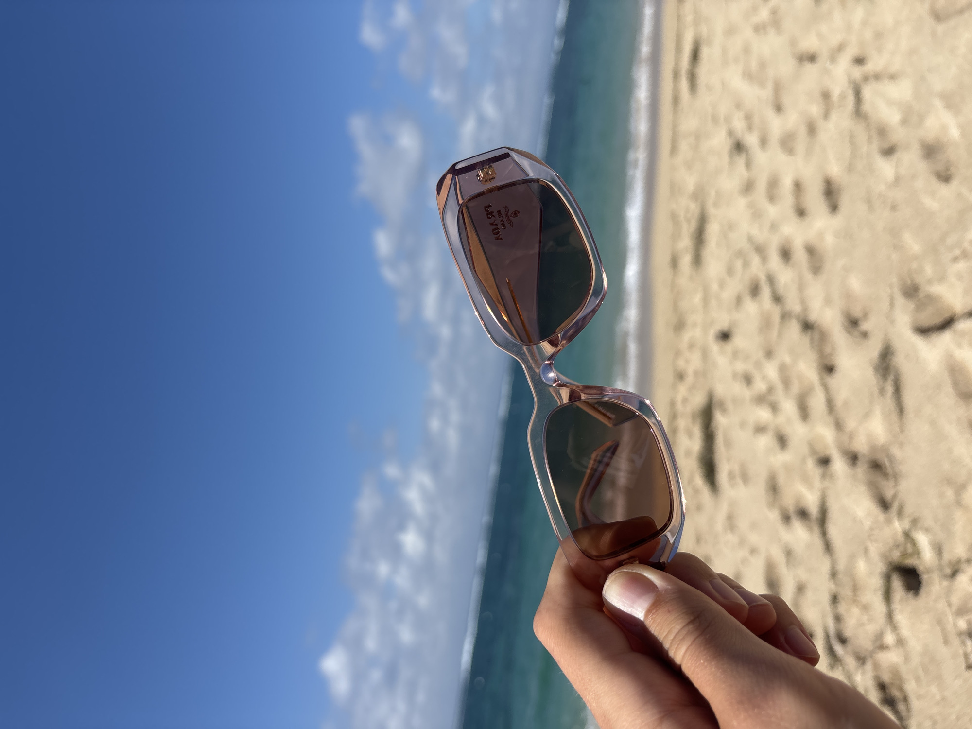 The image presents a close-up, angled shot of a person's hand holding a pair of sunglasses against a scenic beach backdrop. The hand, with fair skin and well-kept nails, holds the sunglasses with the lenses facing away from the viewer.
The sunglasses have a translucent, light-colored frame, possibly light pink or champagne, with a geometric or slightly angular design. The lenses are tinted brown. Inside the right temple of the sunglasses, the brand name "PRADA" is distinctly visible in gold lettering, accompanied by "MILANO" underneath it. Further details like "MADE IN ITALY" are also discernable, albeit less clearly.
The background is split diagonally, showcasing a bright blue sky with scattered white clouds on the upper left, and a sandy beach meeting the clear, turquoise-blue ocean on the lower right. Gentle waves are seen breaking on the shore, creating white foam. The sand is light-colored and appears to have a textured surface.
The setting is a sunny day at a beach, likely in Kuta Selatan, Indonesia, indicated by the vibrant sky, clear ocean, and sandy shore. The overall impression is one of relaxation and enjoying a bright, warm day by the sea.