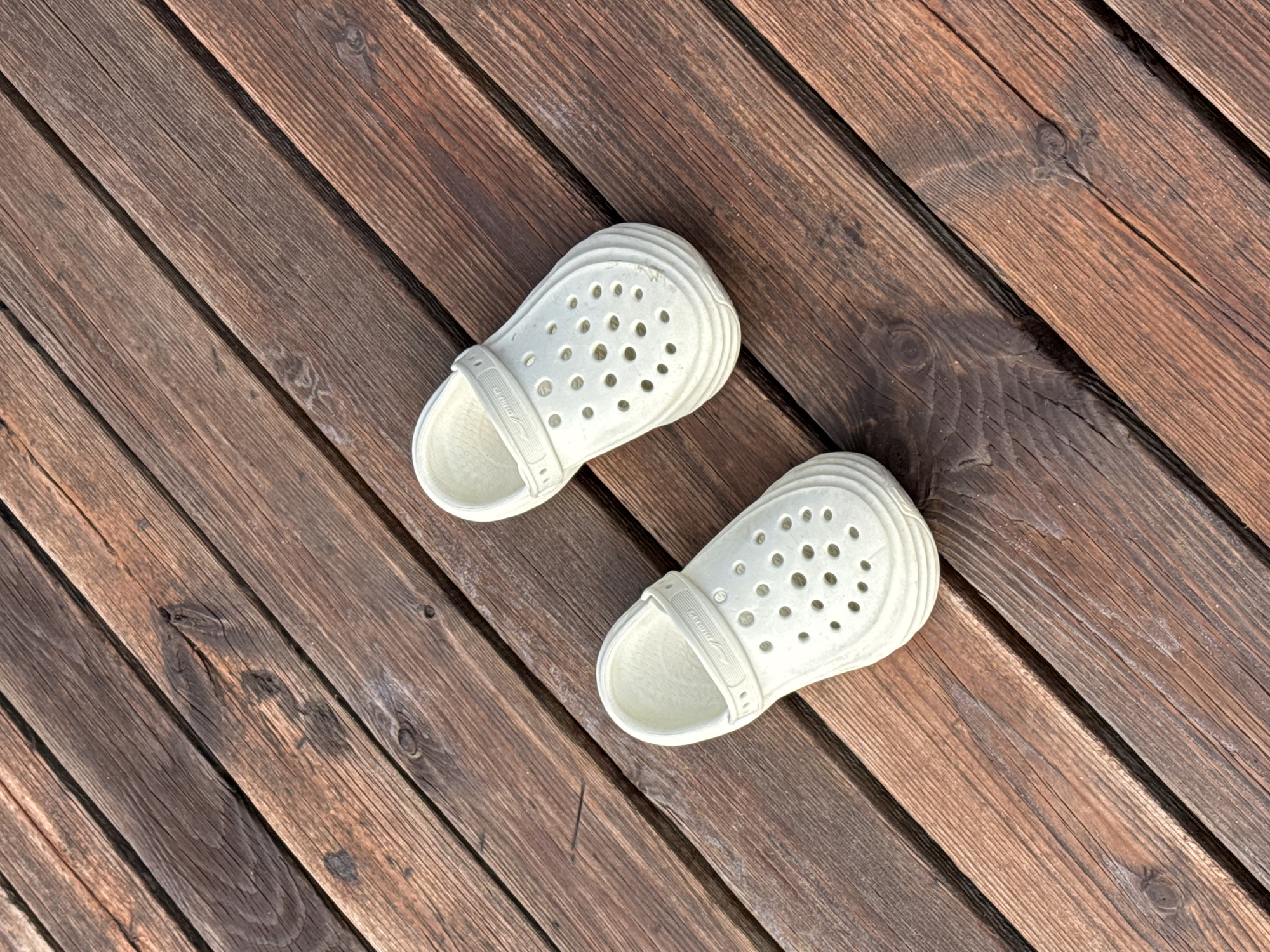 Two white clogs are placed on a wooden deck in Sary-Oy, Kyrgyzstan. The clogs are positioned at an angle, with the left clog slightly closer to the viewer. The wooden deck is made of weathered, brown planks that run diagonally across the image. The planks show visible grain and some knots. The lighting suggests it is daytime, with shadows cast by the clogs.