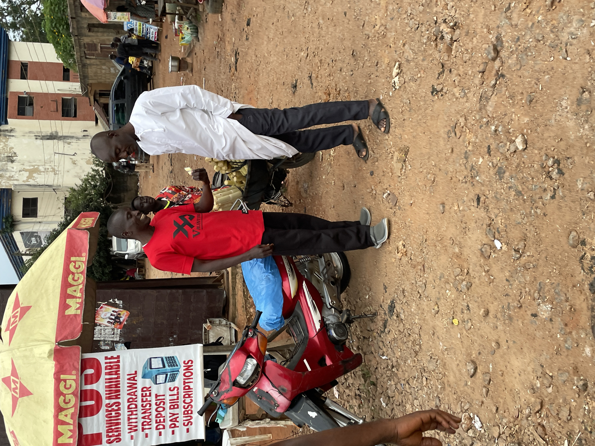 In an outdoor marketplace setting in Jos, Nigeria, two men are engaged in conversation. One man, wearing a red t-shirt and black pants, stands to the left, holding a few stalks of what appears to be sugarcane. The other man, dressed in a white traditional kaftan and black pants, stands to the right, his right hand in his pocket. They are positioned near a red motorcycle covered with a blue tarp and a stall with a yellow and red umbrella displaying "MAGGI" branding. A sign behind the stall lists "P.U.S. SERVICES AVAILABLE - WITHDRAWAL - TRANSFER - DEPOSIT - PAY BILLS - SUBSCRIPTIONS." The ground is a mixture of dirt and small stones, with debris scattered around. In the background, other market stalls and buildings are visible, along with a few parked vehicles and other individuals moving about the marketplace. The overall atmosphere suggests a busy commercial area.