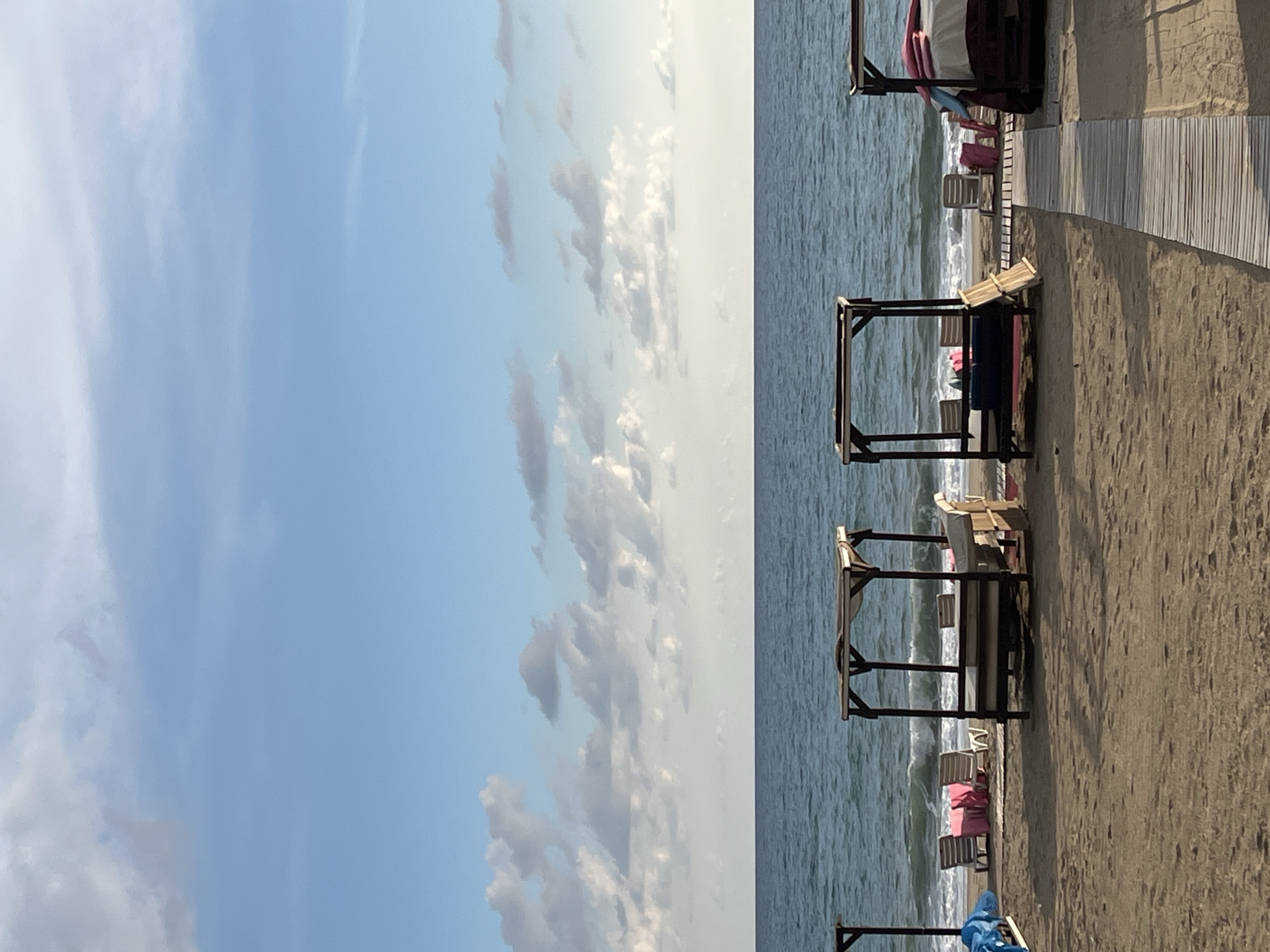 The image captures a beach scene in Alanya, Turkey. The sky is blue with scattered white clouds. The sea is calm and stretches to the horizon. Along the beach, there are several sun shelters and beach chairs. A wooden boardwalk runs parallel to the shoreline. There are no people visible in the frame. The sand appears light brown.