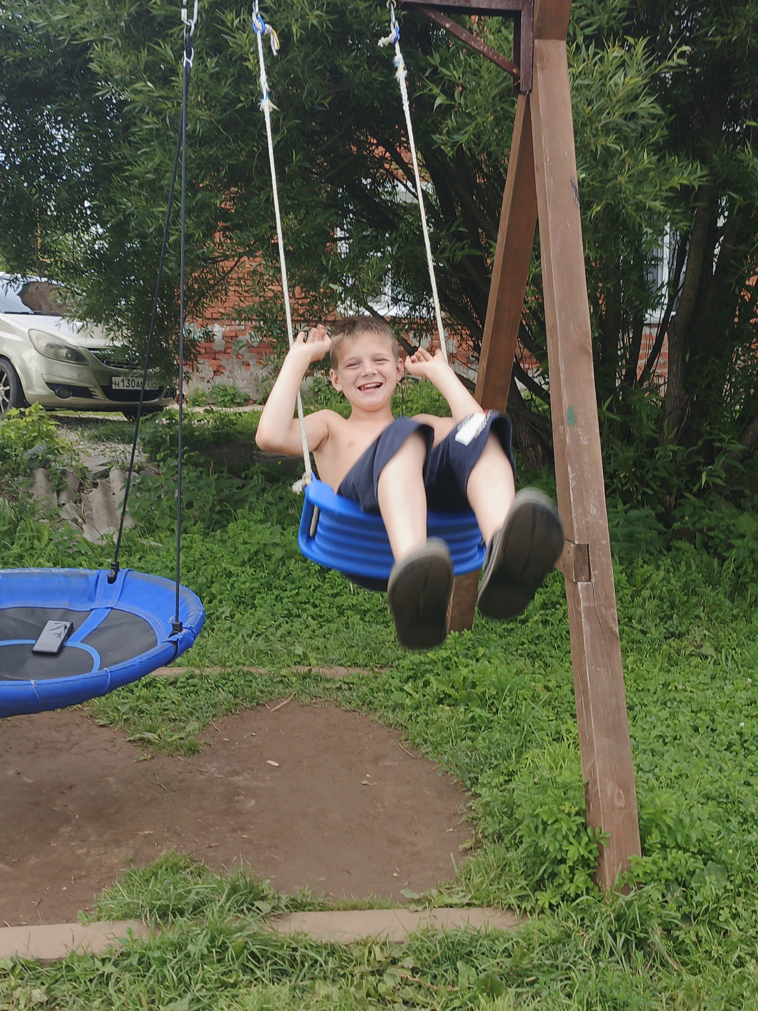 In Medvedyovka, Russia, a young boy is captured mid-swing on a blue plastic swing seat. He is shirtless, wearing dark blue shorts, and appears to be laughing with his arms raised. The swing is attached to a wooden frame structure. To the left of the swing, a blue circular nest swing hangs. The background consists of lush green foliage, a brick building, and a silver car parked on an elevated area. The ground beneath the swings is a mix of dirt and grass.
