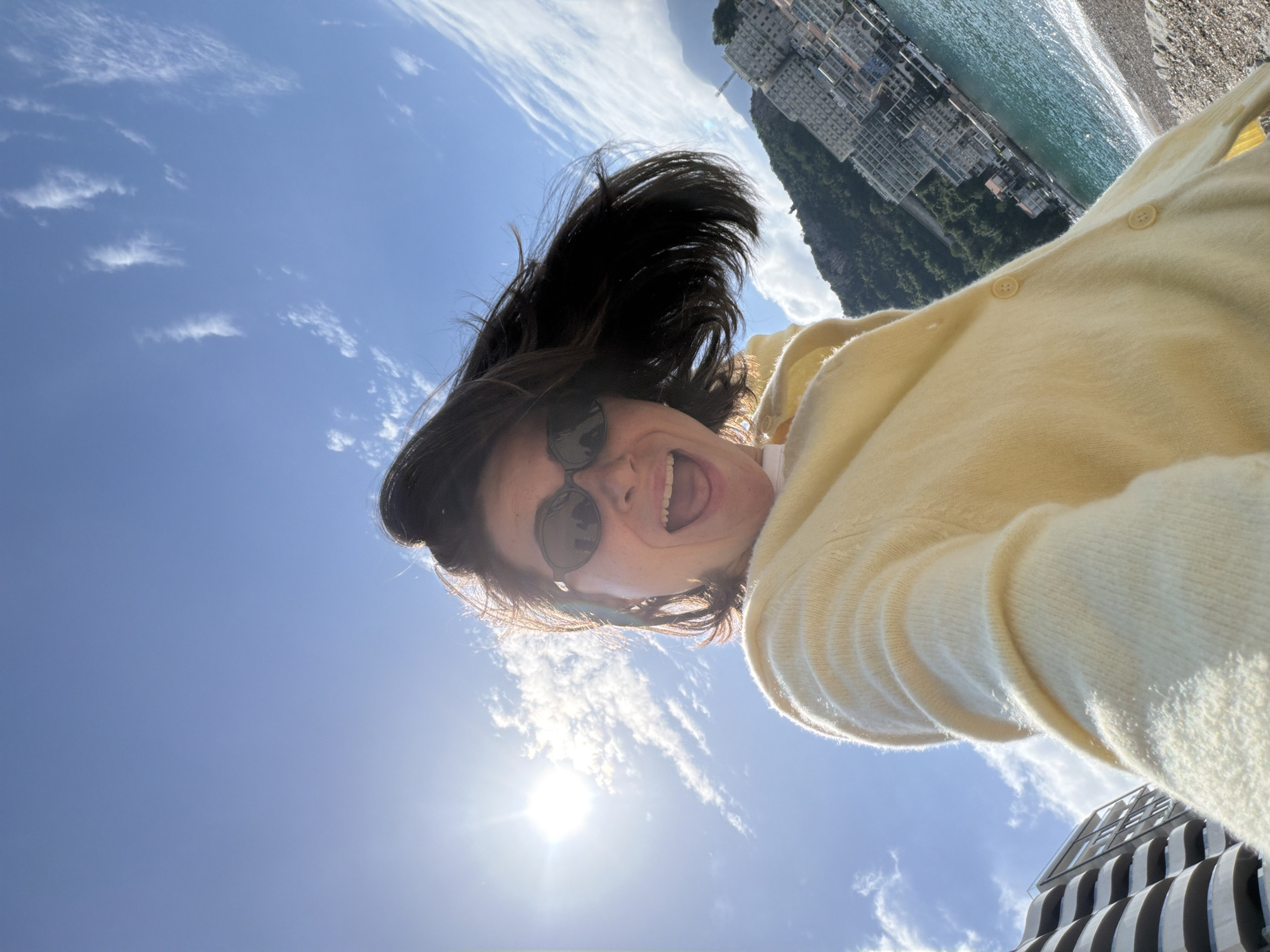 A selfie photograph looking upwards towards the sun. The subject is a person wearing sunglasses and a yellow cardigan, with dark hair blowing upwards. The person is sticking their tongue out and smiling. The background features a blue sky with scattered white clouds, a building complex on a hillside, and the sea and beach. The location appears to be Rafailovići, Montenegro, based on the distinctive building style and coastal features.