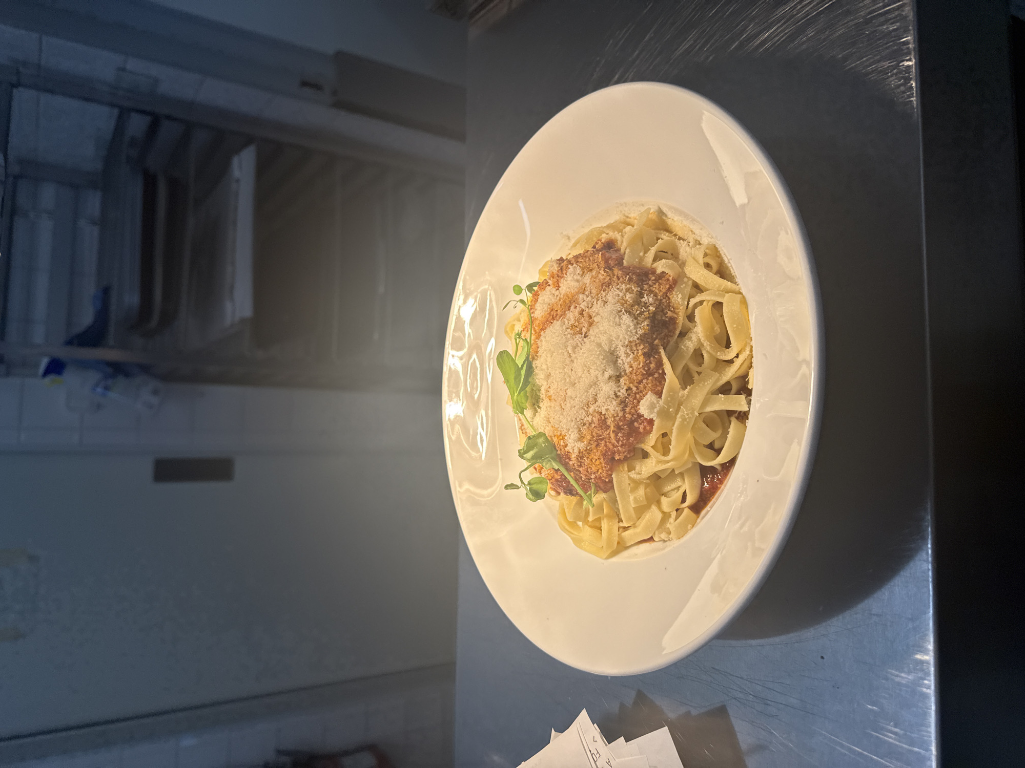 A white plate of fettuccine pasta with bolognese sauce and grated parmesan cheese is displayed. The pasta is topped with a rich red sauce and a generous sprinkle of cheese. A few sprigs of fresh green herbs add a touch of color. The plate is set on a dark gray surface with a metallic sheen, possibly a countertop or table, and is illuminated by soft lighting. In the background, out of focus, is a kitchen or service area with stainless steel appliances and tiled walls. The overall scene suggests a prepared meal ready to be served or enjoyed. There is no visible text. The context indicates this is in Rivesaltes, France.