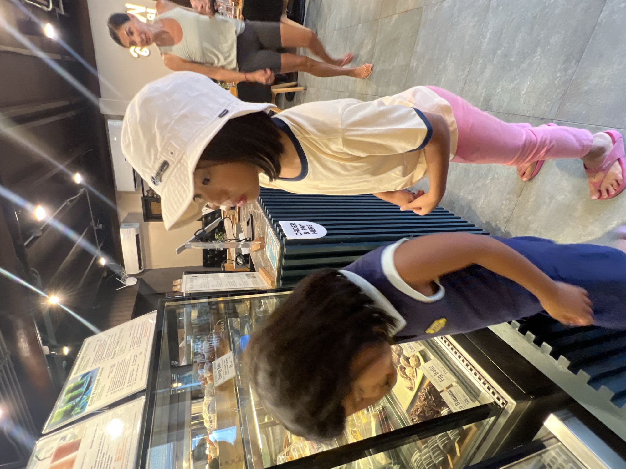 The image, taken with a tilted perspective, captures a scene inside a cafe or bakery in Baan Lamai, Thailand. Two young children are prominently featured in the foreground and midground, both engrossed in looking at items displayed in a glass counter. The child closest to the camera has dark hair and wears a blue and white sleeveless top. Behind them, another child wears a white bucket hat, a light yellow t-shirt with blue trim, pink pants, and pink flip-flops.
In the background, an adult in a light tank top and dark shorts stands observing the children, partially obscured. The setting features modern grey tiled flooring, a dark ceiling with exposed track lighting, and bright artificial illumination.
The glass display cases hold various baked goods and treats. Menu boards are visible on the counter, with text indicating offerings like "Cold Pressed Juice" and "Energy Balls." Specific items visible and their prices include "Fig Balls" at "115" and "Co Banana" at "125," along with "Cookie." A clear sign on the counter instructs customers to "ORDER & PAY HERE." A "DELL" logo is visible on a monitor behind the counter, and a partial letter "N" from a larger sign is also present. The children appear focused on their choices, creating an impression of anticipation at the cafe.