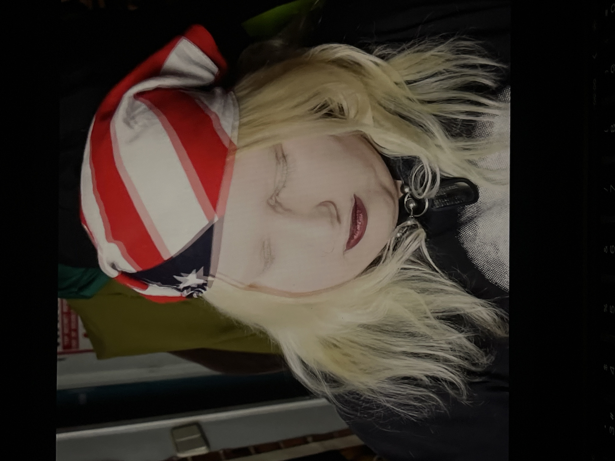 A close-up portrait, oriented diagonally, features a person with light blonde hair and notably pale skin, possibly wearing makeup. Their eyes are closed, and red lipstick is visible on their lips. They are wearing a red and white striped hat or bandana that covers part of their head, featuring a blue section with a single white star, similar to design elements found in the U.S. flag. Around their neck, a black choker or collar with metallic rings is visible. The person is dressed in a dark garment, possibly a black top or jacket, with a subtle textured pattern visible on the right side.
The background is dark and out of focus, revealing hints of green and olive-colored fabric elements behind the person's head, and a light horizontal surface below. The subdued lighting suggests an indoor setting. The person appears to be resting or sleeping due to their closed eyes and calm facial expression. No specific activity or event is clearly depicted beyond this state of repose. There are no direct visual cues within the image to indicate the location as Torrent, Spain. Faint, blurred numerical and symbolic characters (such as '0', '9', '8', '%', '4', '#') are visible along the far right edge of the frame, possibly from an overlaid digital interface.