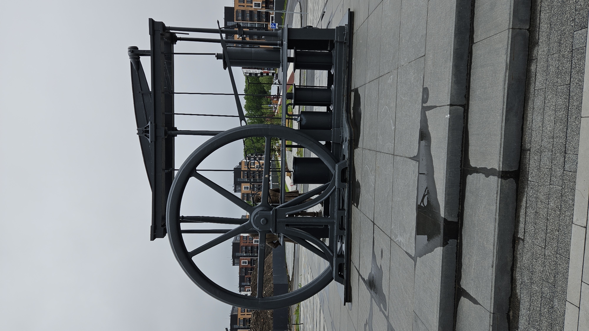 A piece of equipment, potentially a pump or a wheel and gear mechanism, stands on a paved surface in an urban area. The equipment is predominantly gray in color, with a large wheel featuring a series of spokes as a prominent feature. Behind the equipment, there are buildings, likely residential, suggesting an urban or suburban location. The sky is overcast, and there are areas on the pavement that appear wet, indicating recent rainfall or damp conditions.