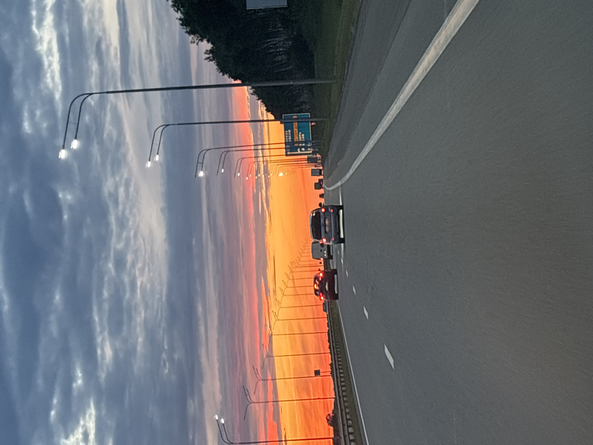 The media file is a vertical, slightly tilted, wide-angle photograph taken from the perspective of a moving vehicle on a highway. The scene is dominated by a dramatic sunset sky, transitioning from deep blue and grey clouds on the left to vibrant orange and pink hues towards the right. A line of streetlights with illuminated bulbs stretches along the left side of the highway, silhouetted against the colorful sky.
The highway itself is paved and appears to have multiple lanes, with white lane markings visible. Several vehicles are traveling on the highway, including cars and what appears to be a van or larger vehicle. The streetlights suggest this is a well-maintained road, possibly an expressway or a major highway.
On the right side of the highway, a line of trees forms a dark backdrop against the sunset. A blue road sign with white text is visible in the distance, indicating directions and distances. Though the text is partially obscured and difficult to read clearly due to the distance and angle, some Cyrillic letters can be discerned, suggesting a location in Russia.
The time of day is clearly sunset, indicated by the intense colors in the sky. The weather appears clear enough to allow for a vibrant sunset. The overall mood is serene and picturesque, capturing the beauty of a sunset on a highway journey. The presence of vehicles suggests a normal flow of traffic. There is no clear indication of people within the frame. The location context provided, Bogorodskoe, Russia, aligns with the visible Cyrillic text on the road sign.